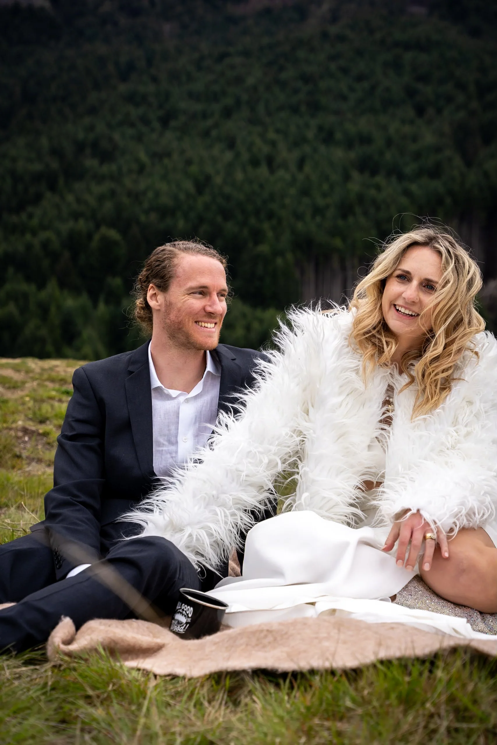 A man and woman sitting on a blanket outdoors, smiling, with a forested mountain background. The woman is wearing a white furry coat and the man is in a dark suit.