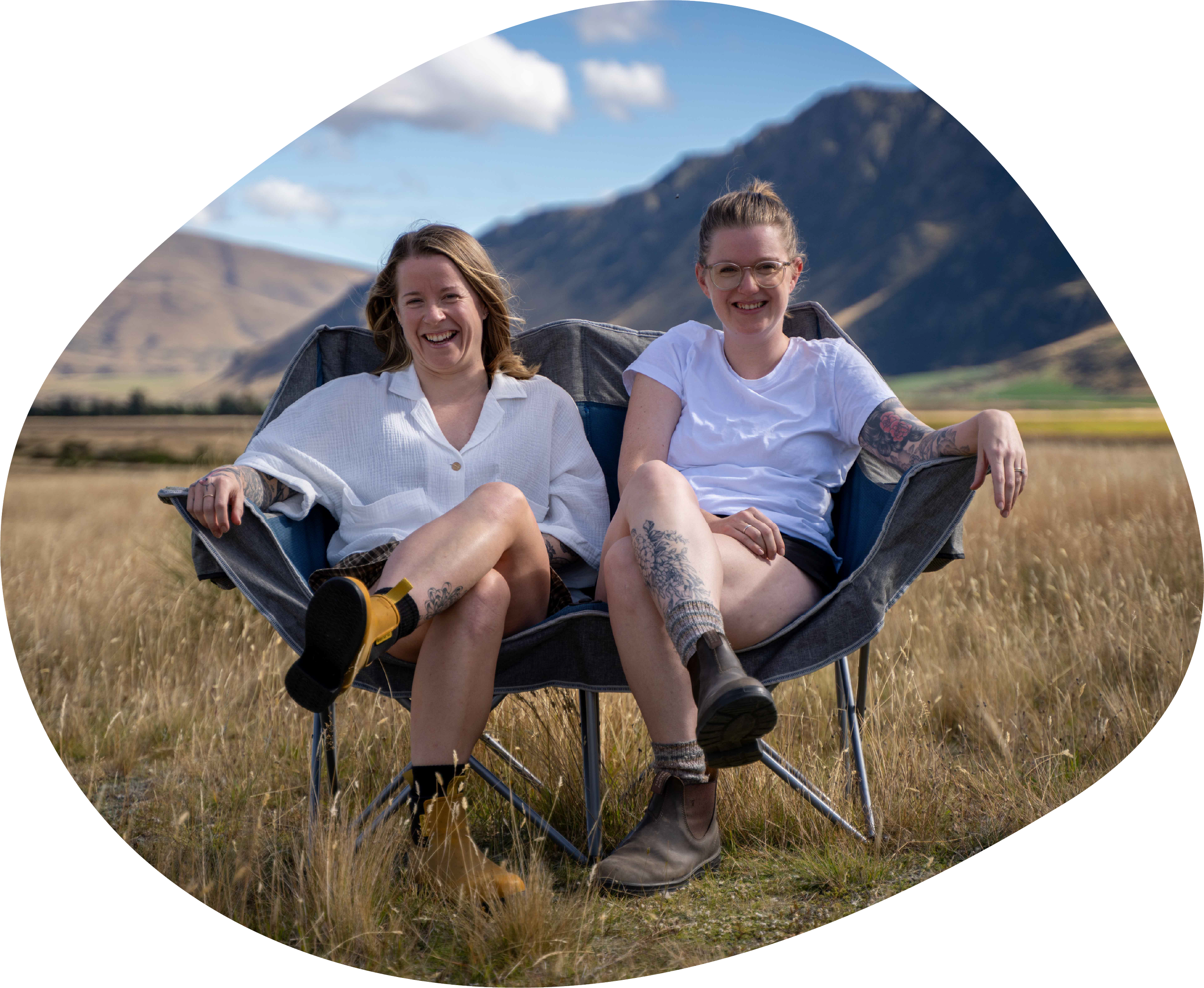 Two women sitting in a portable hammock chair outdoors in a grassy field, smiling at the camera with mountains and a partly cloudy blue sky in the background.