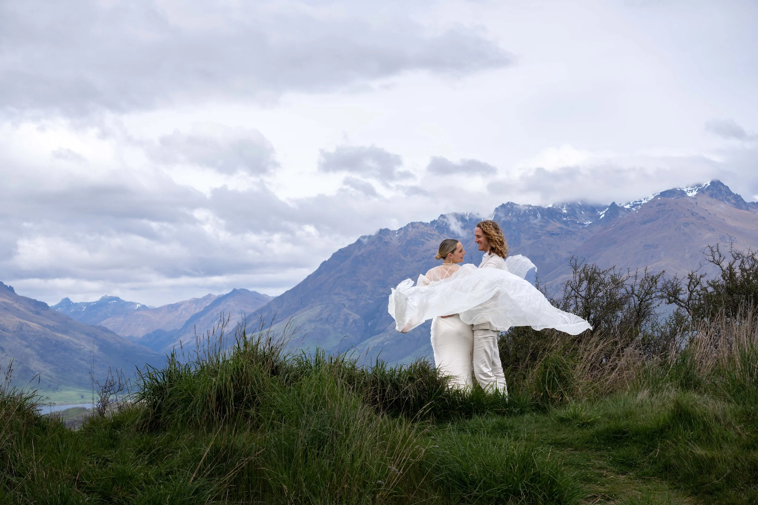 A couple in wedding attire standing in a grassy field with mountains and cloudy sky in the background.