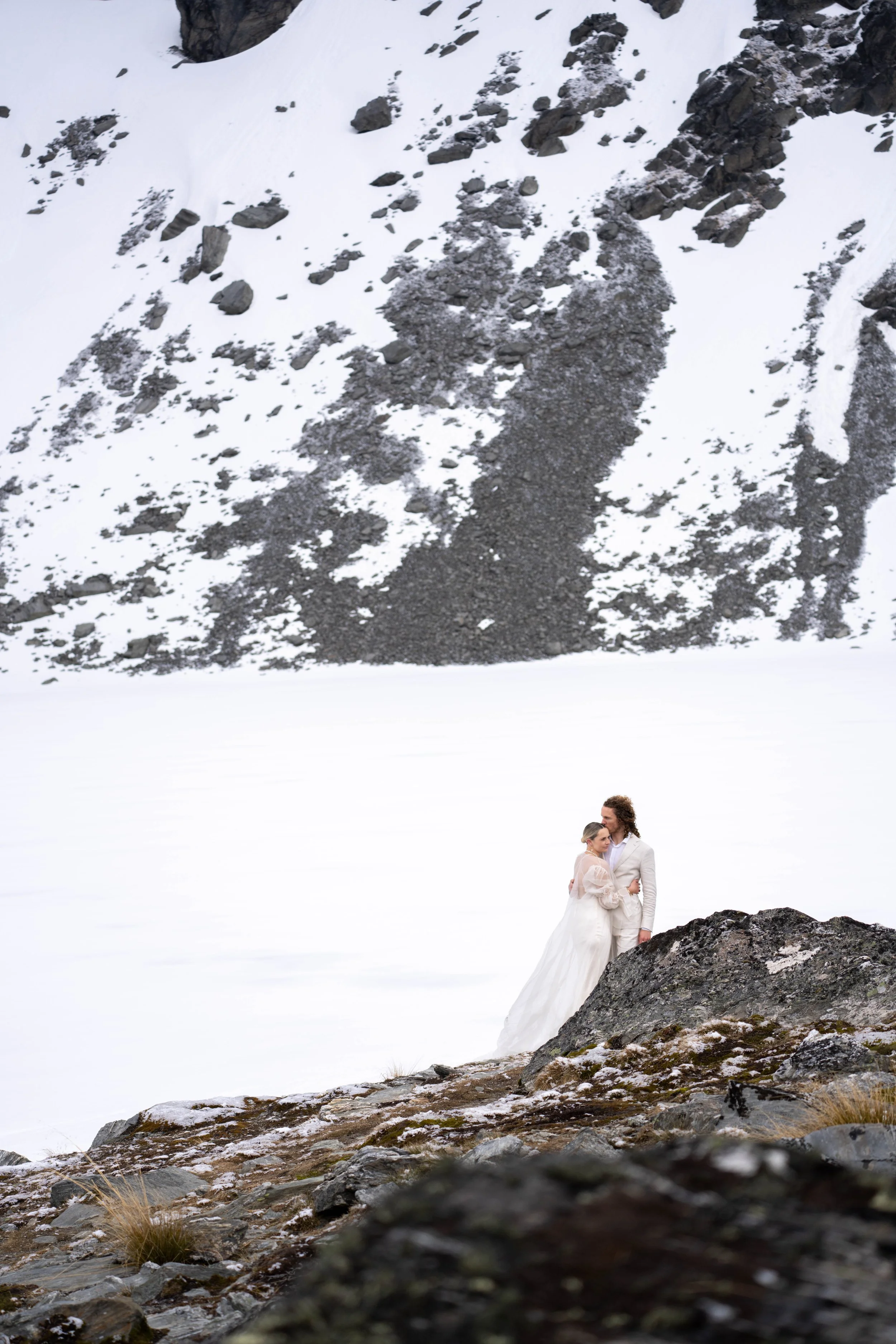 A bride and groom dressed in wedding attire standing closely together outdoors in a snowy mountainous landscape.
