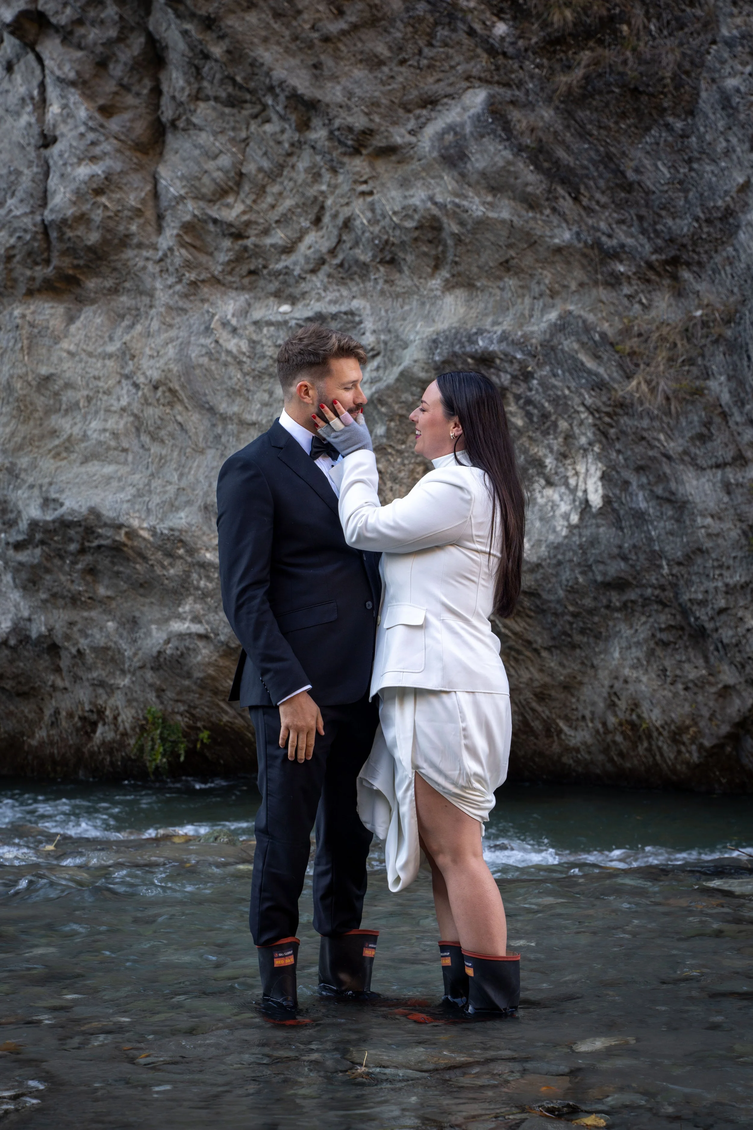 A couple in formal attire standing in a shallow creek with a rocky background. The woman is touching the man's face while smiling, and both are wearing waterproof boots.