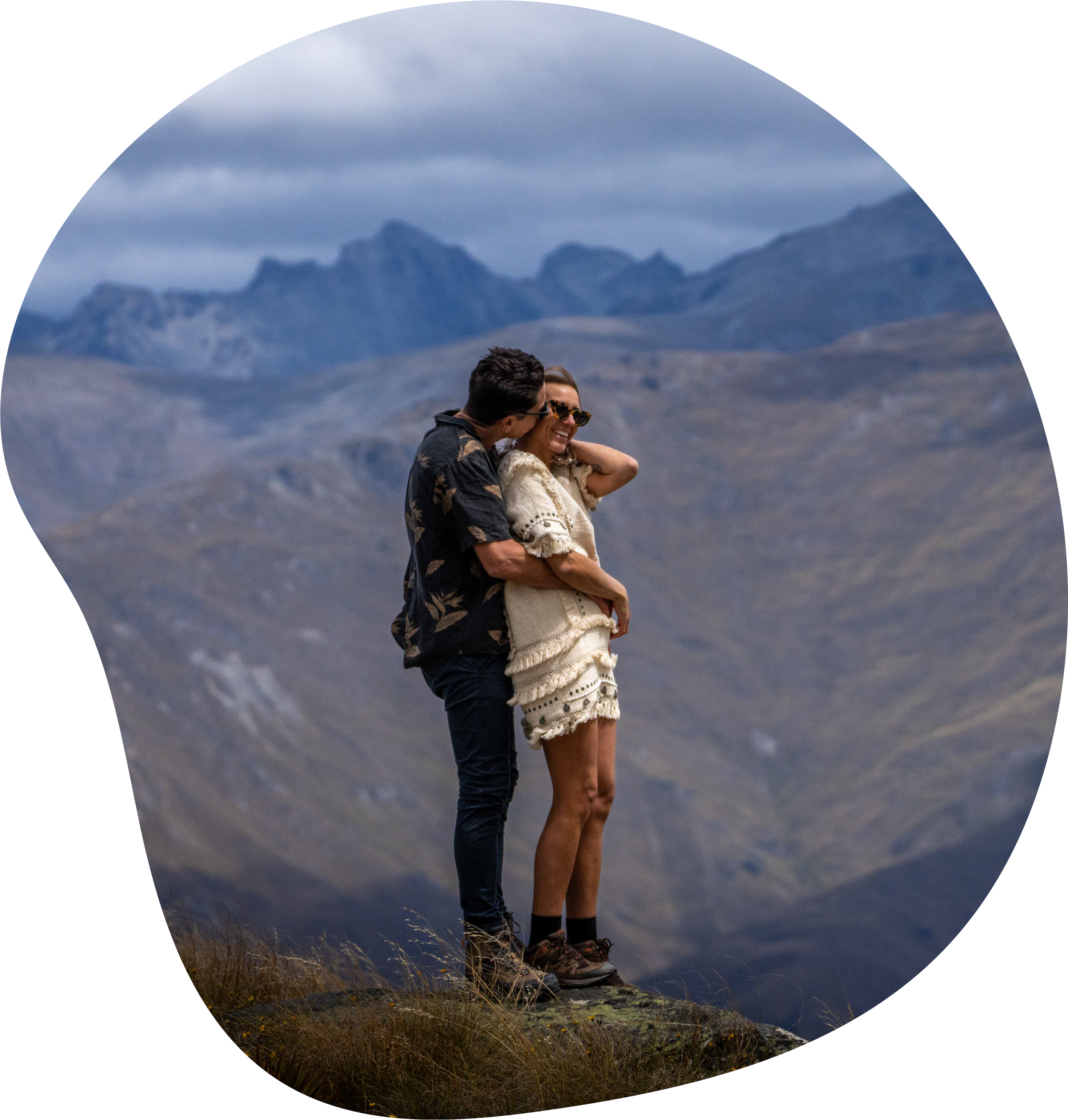A couple standing on a rock in a mountainous landscape with cloudy skies, with the man kissing the woman on the cheek, both smiling.