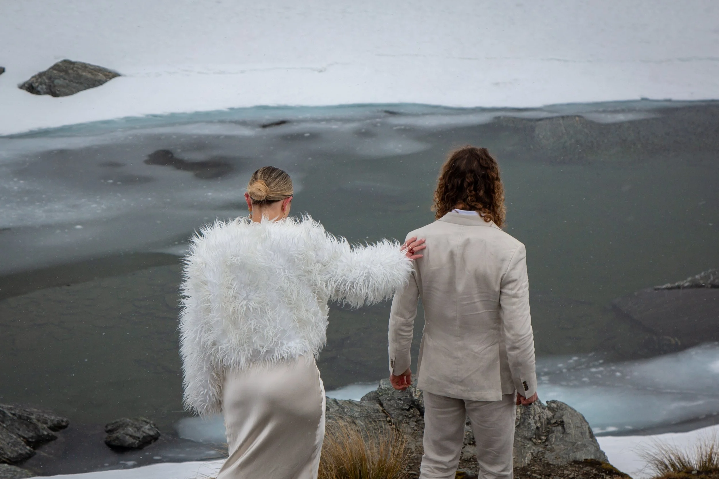 Two people dressed in light-colored clothing stand on rocky terrain near a body of water with snow and ice, one person touching the other's shoulder.