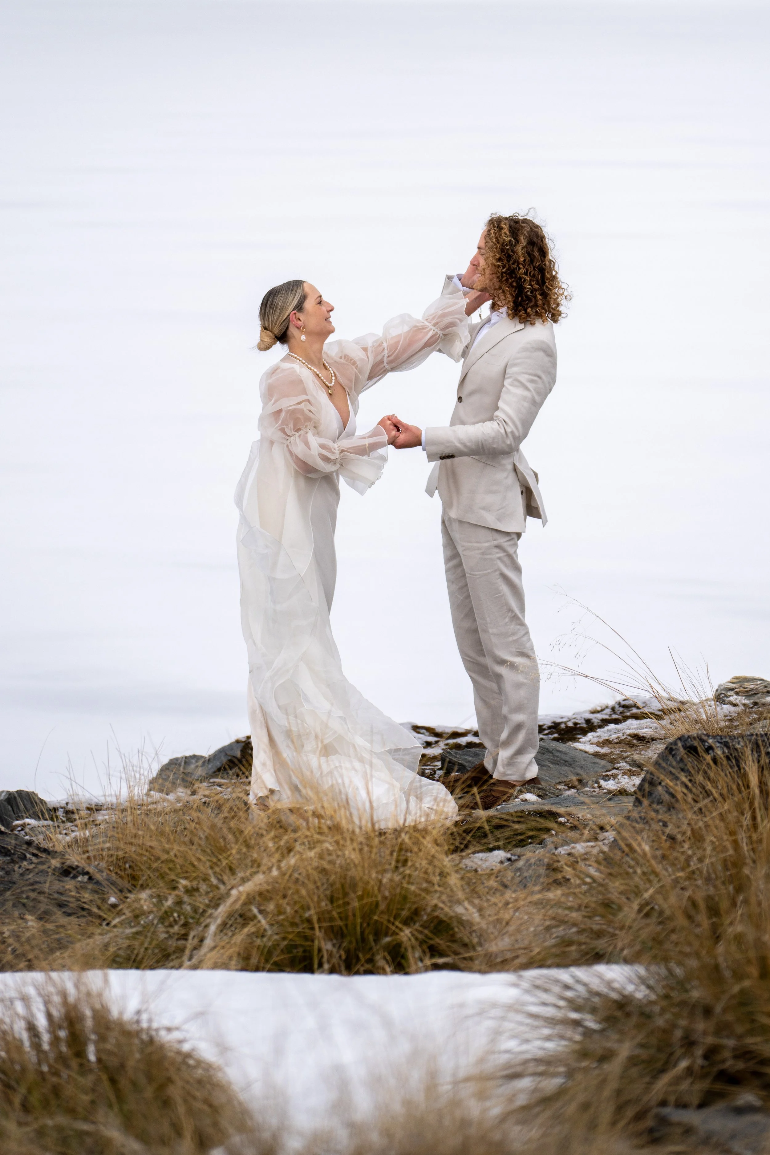 A bride and groom standing outdoors on rocky terrain with grasses, holding hands and gazing at each other, with a plain white background.