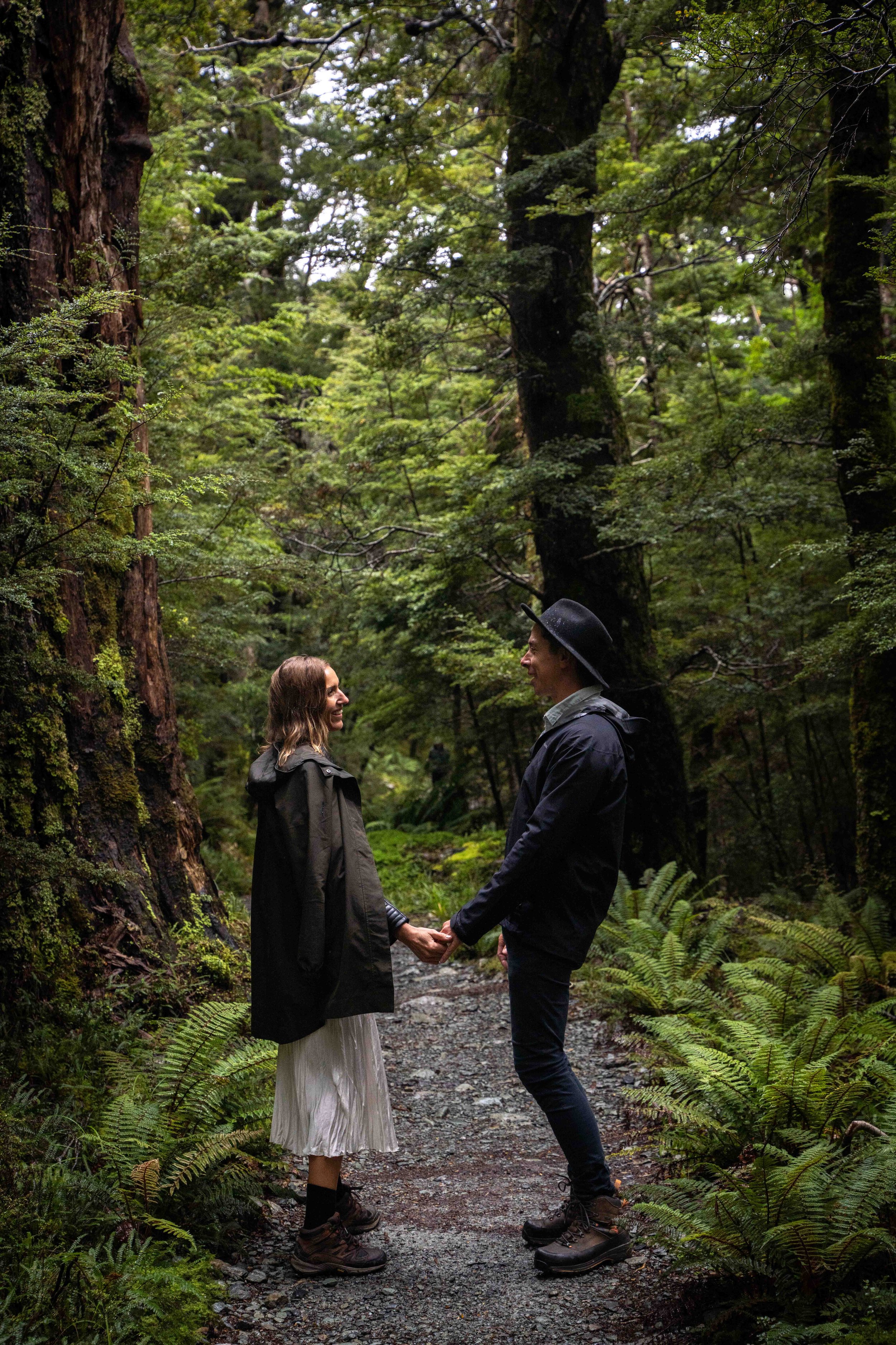 A couple holds hands and smiles at each other on a forest trail, surrounded by tall trees and green foliage.
