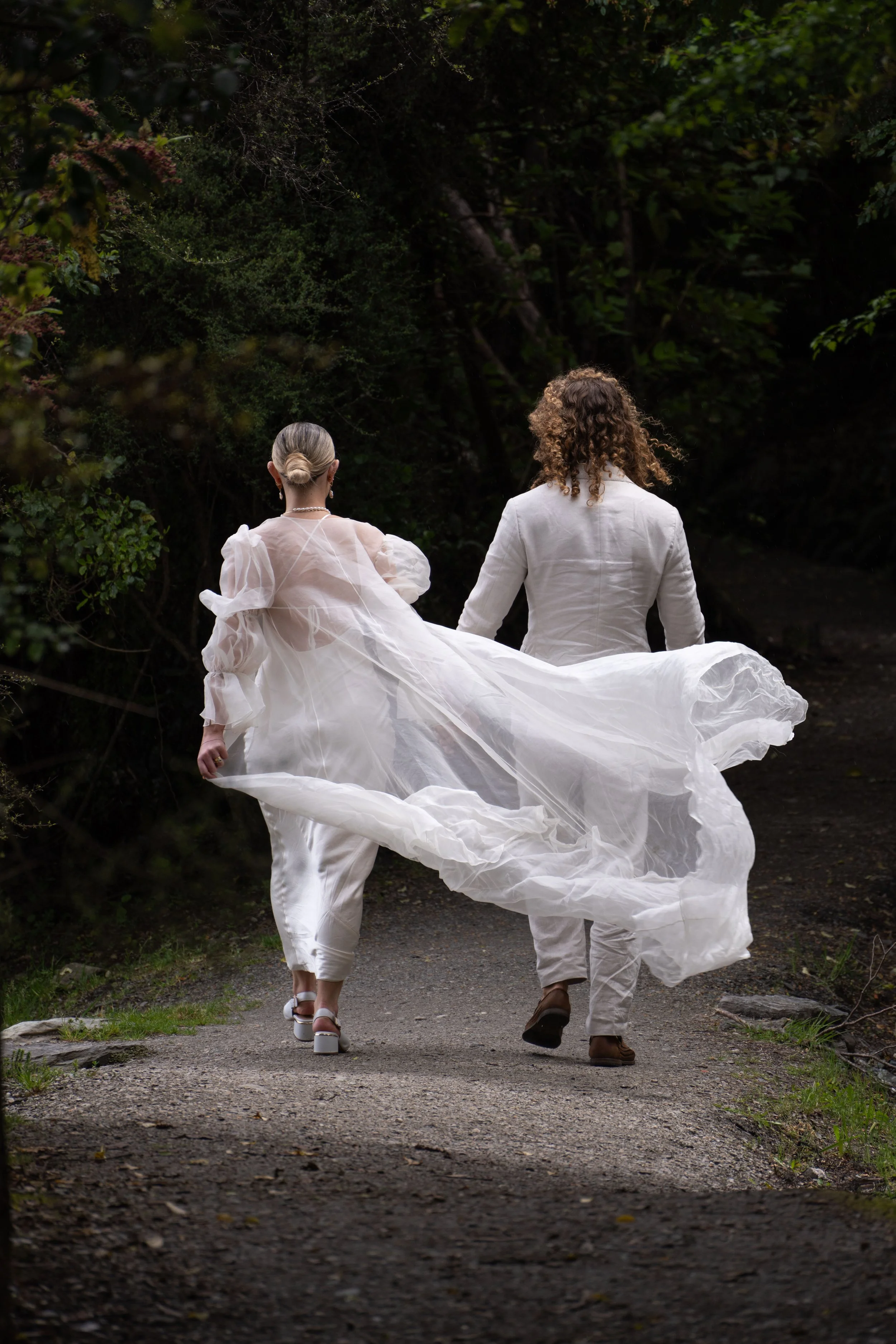 A person in a flowing white dress and another in a white suit walk away down a wooded dirt path.