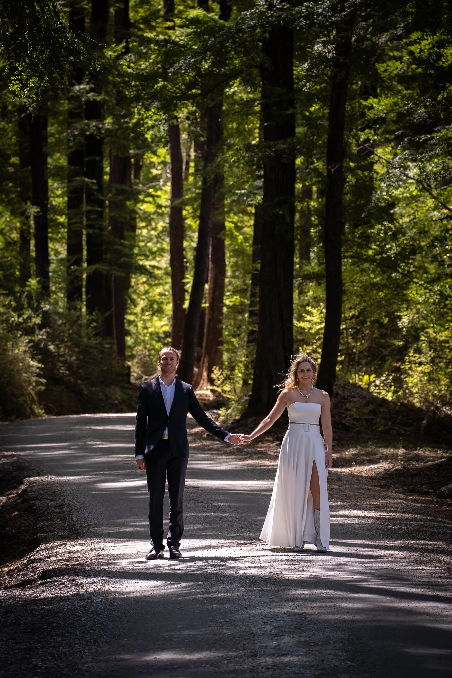 A couple in wedding attire walking hand in hand on a forested dirt road with tall trees and green foliage.