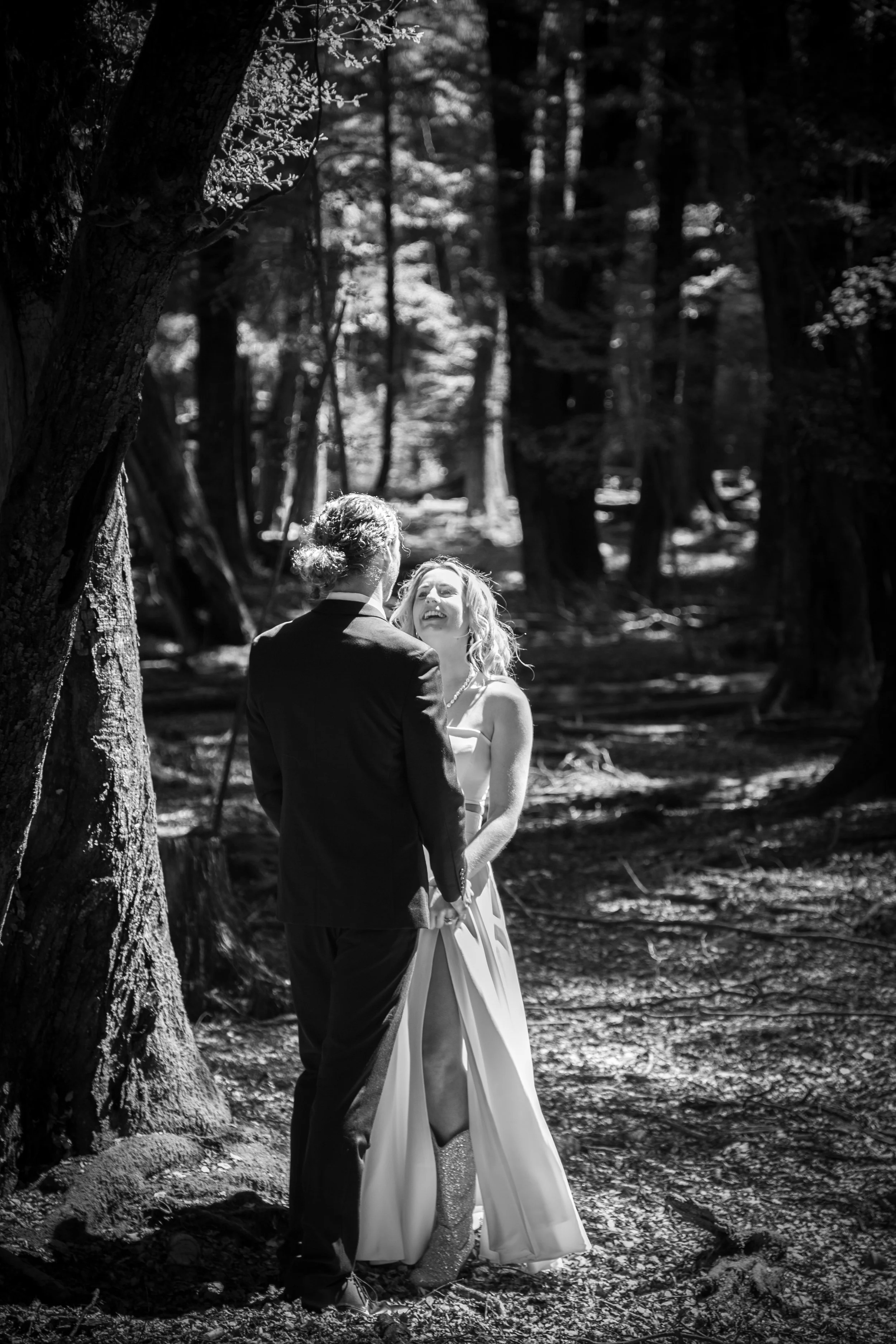 A bride and groom holding hands and smiling in a wooded forest during a wedding photo shoot in black and white.
