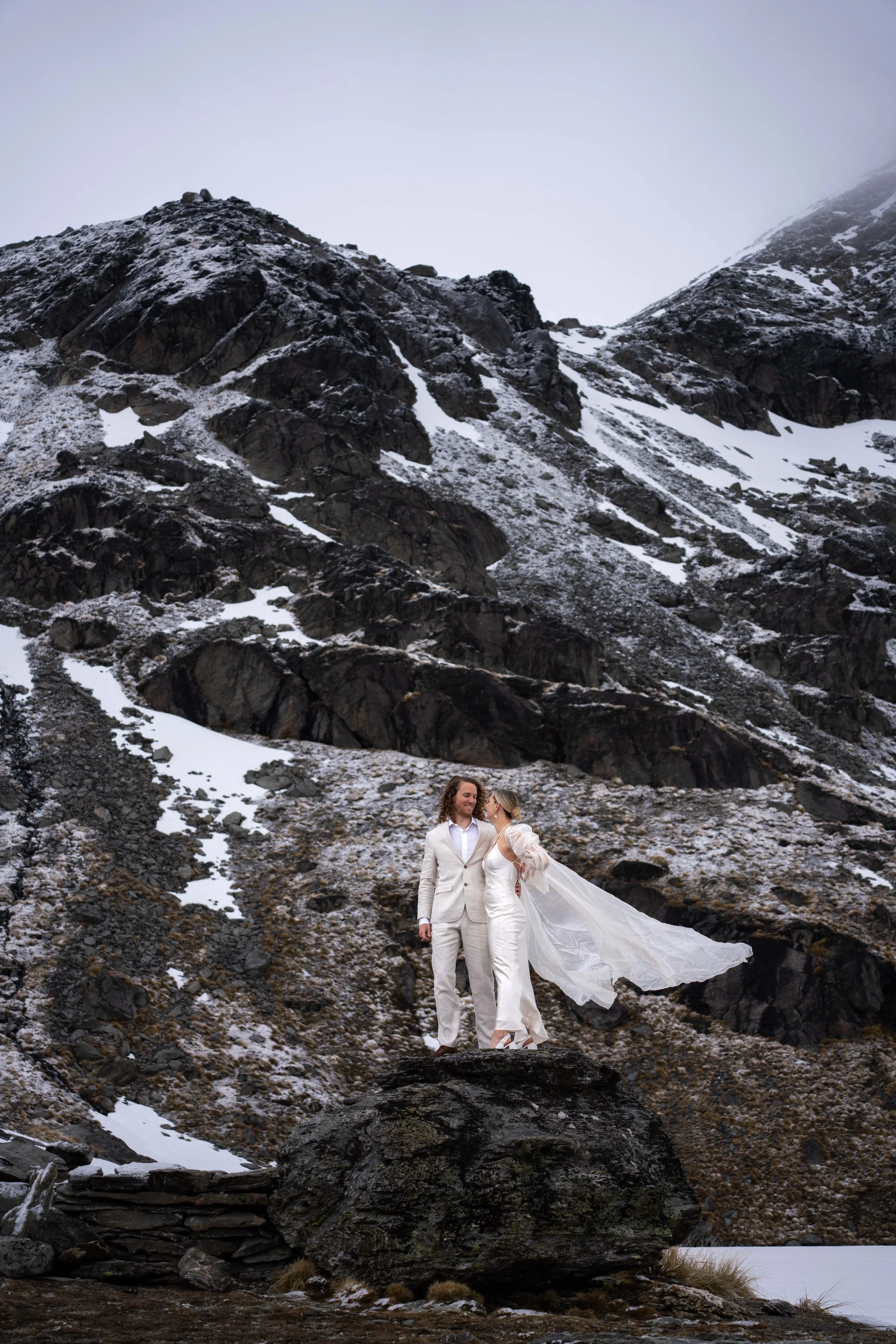 A bride and groom standing on a large rock in a mountainous, snowy landscape. The bride's veil flows in the wind, and they are dressed in white wedding attire.