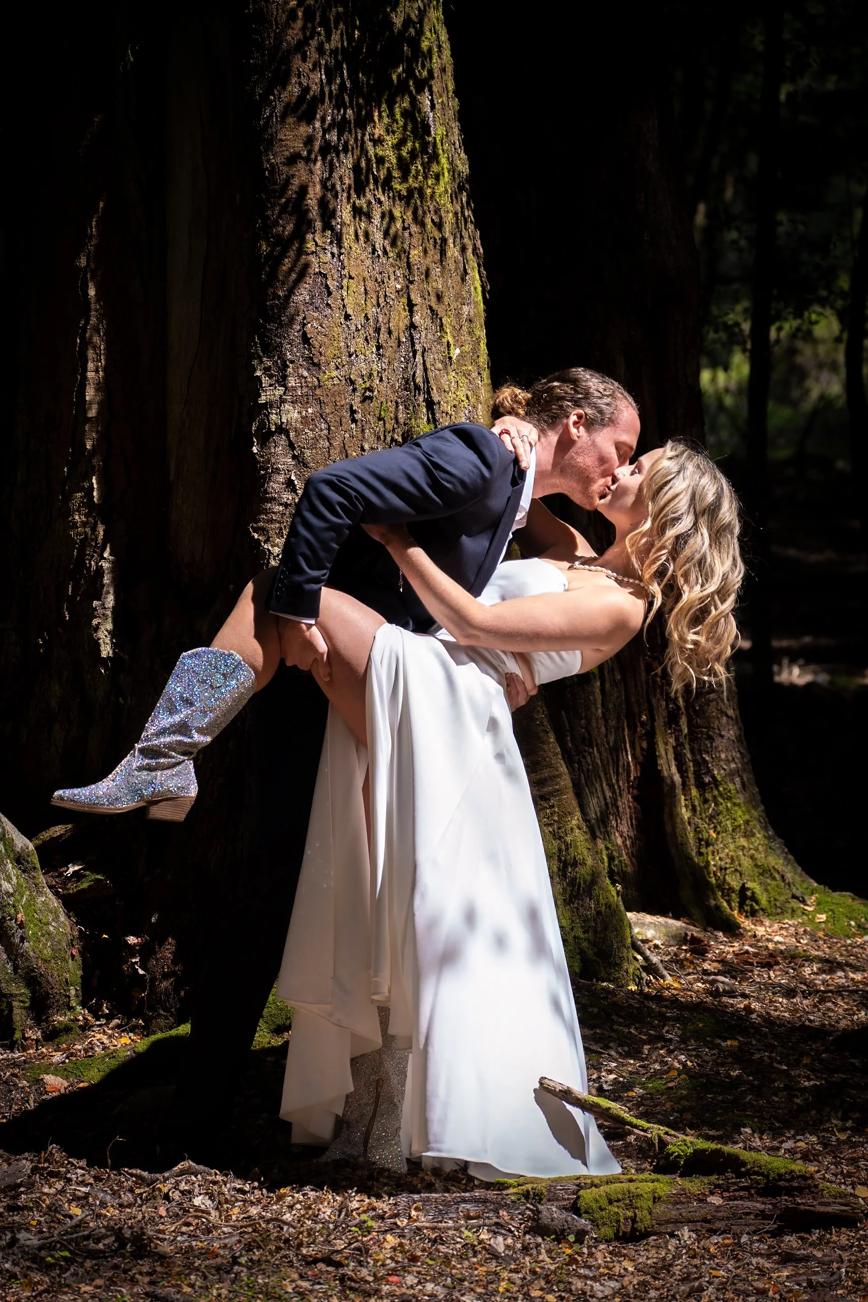 A couple in wedding attire sharing a kiss in a forest, with the groom holding the bride and she wearing sparkly boots.