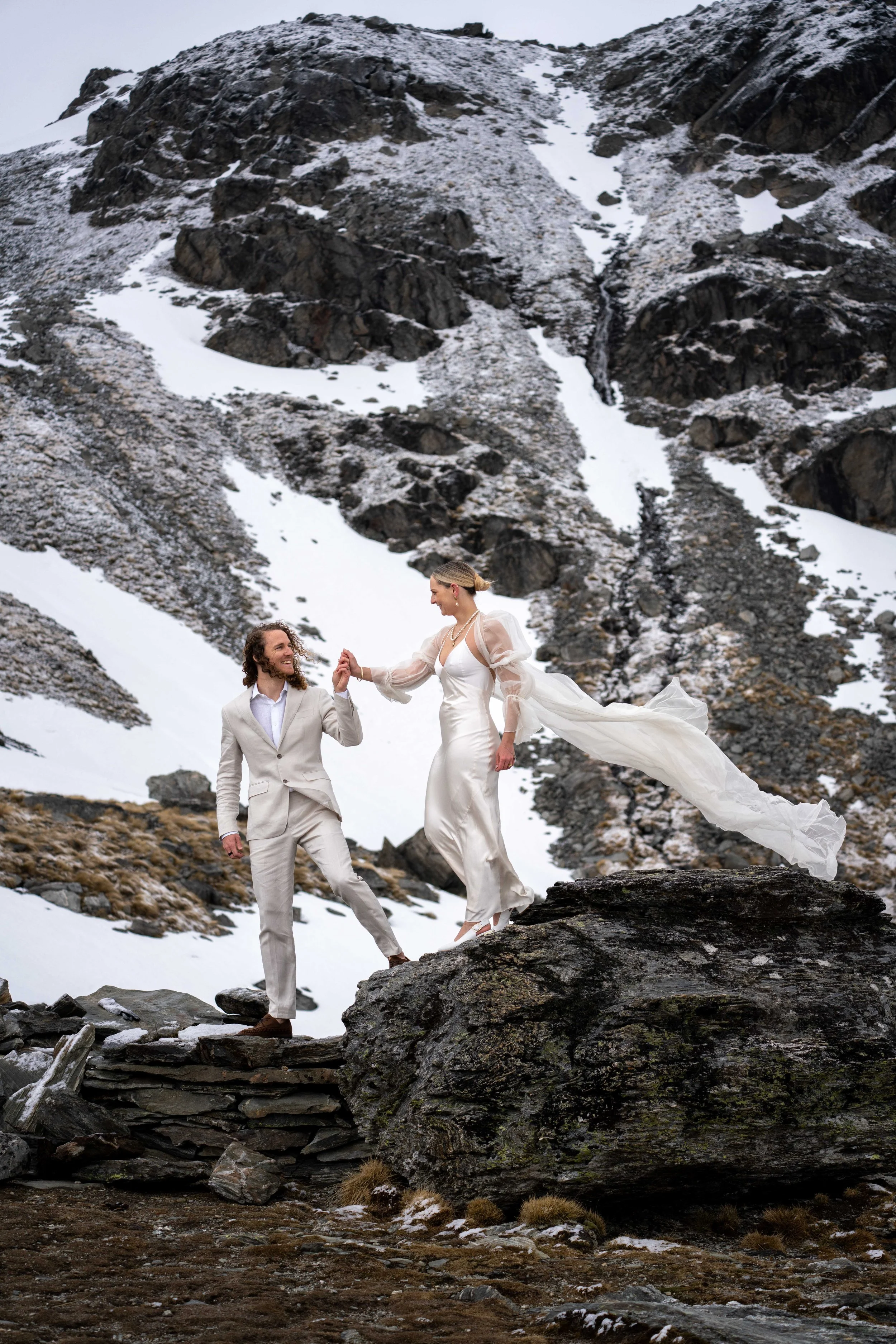 A bride and groom in wedding attire holding hands on a rocky mountain landscape with snow patches.