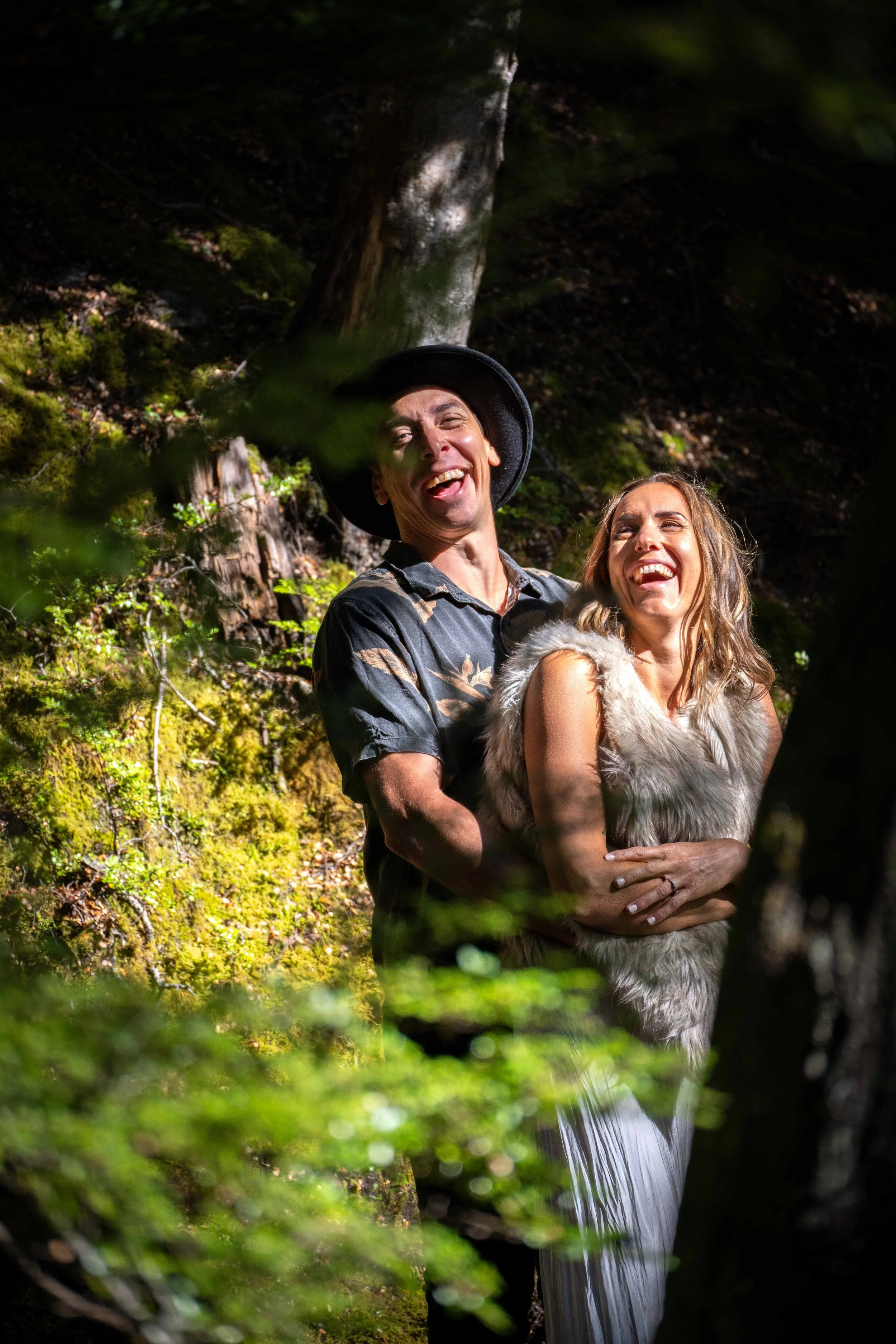 A man and woman standing outdoors in a forest, smiling and laughing, with the man wearing a hat and a patterned shirt, and the woman dressed in a fur vest and a long white skirt.