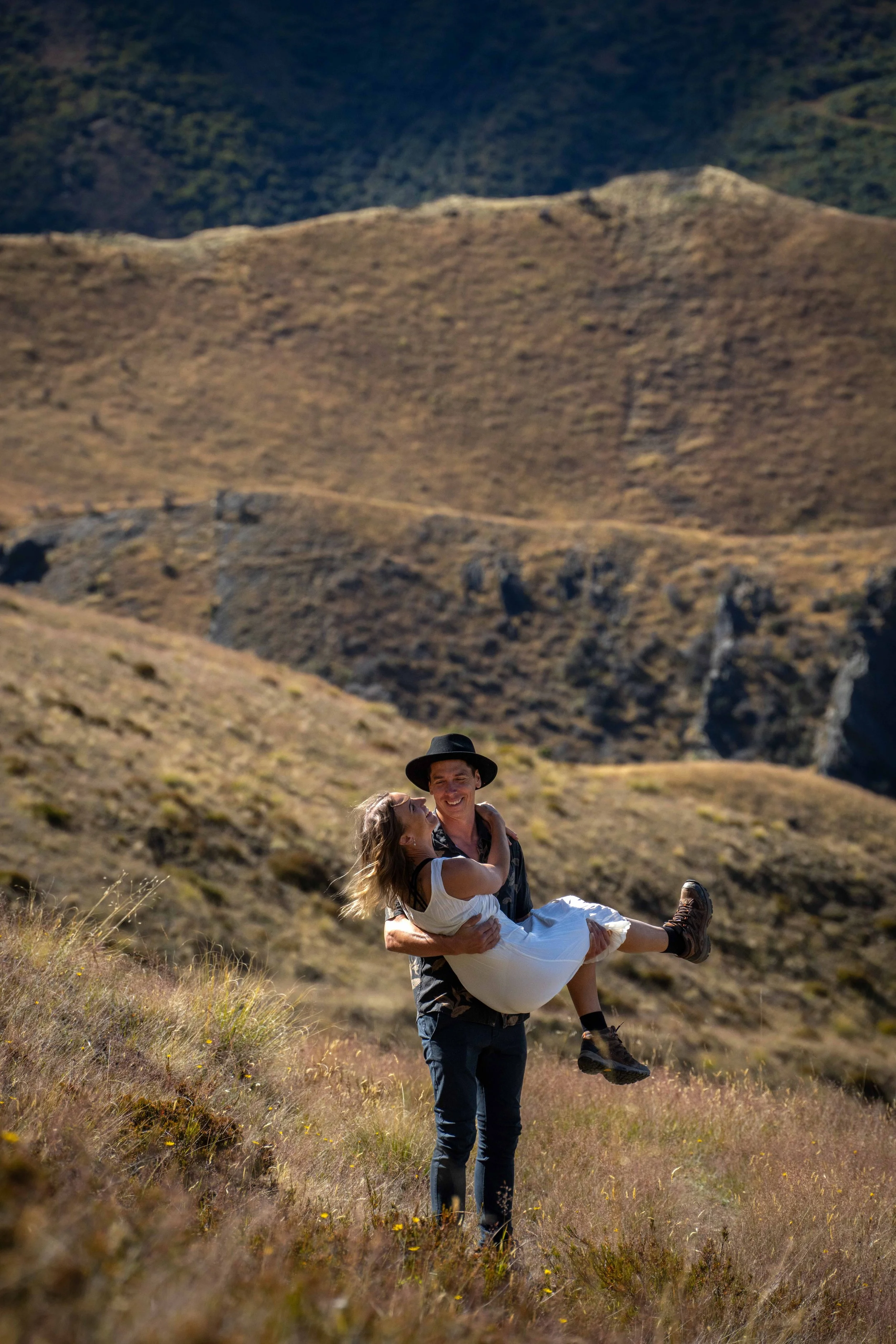 A man in a black hat and patterned shirt carries a woman in a white dress in a hilly landscape with dry grass and rocky cliffs in the background.