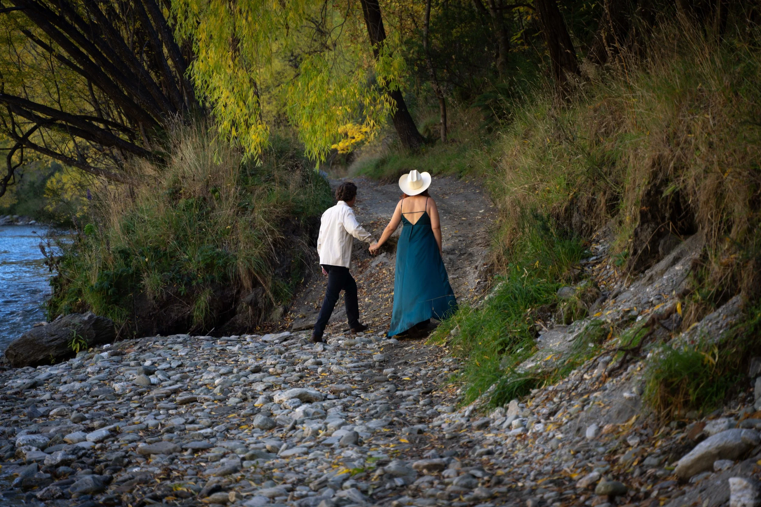 A man and woman holding hands walk along a rocky trail beside a body of water, surrounded by trees with green and yellow leaves.