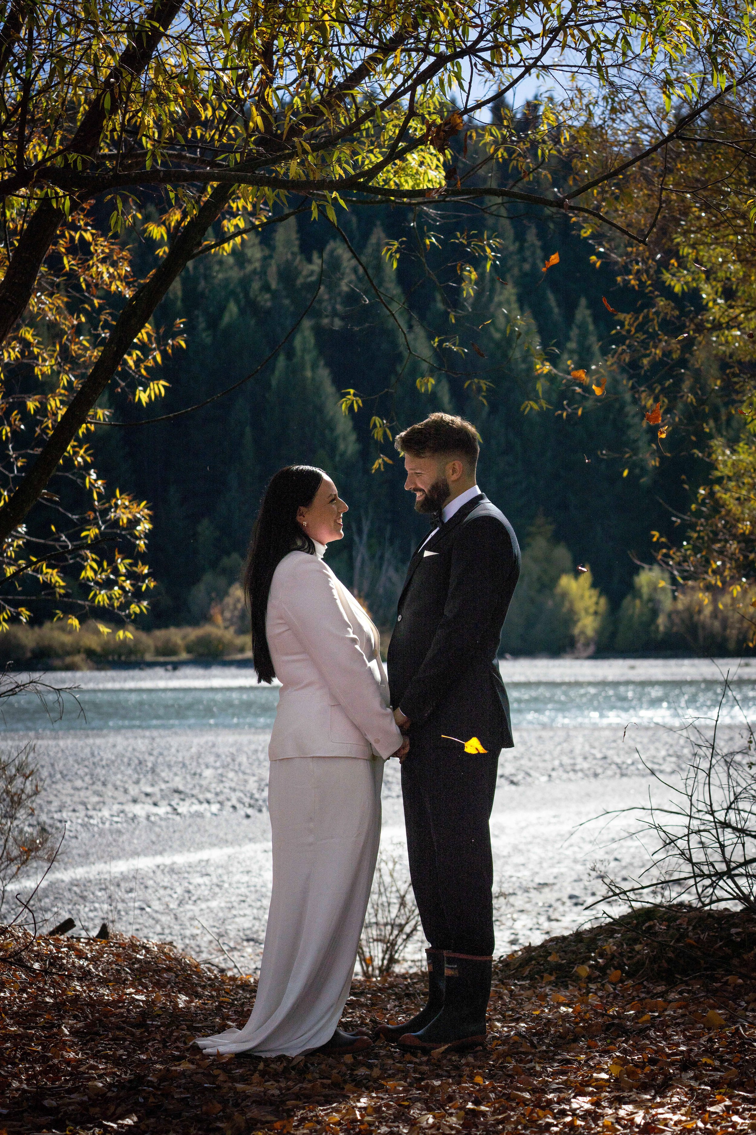 A couple dressed in formal wedding attire, standing on a forest floor covered with fallen leaves, holding hands and facing each other with a river and trees in the background.