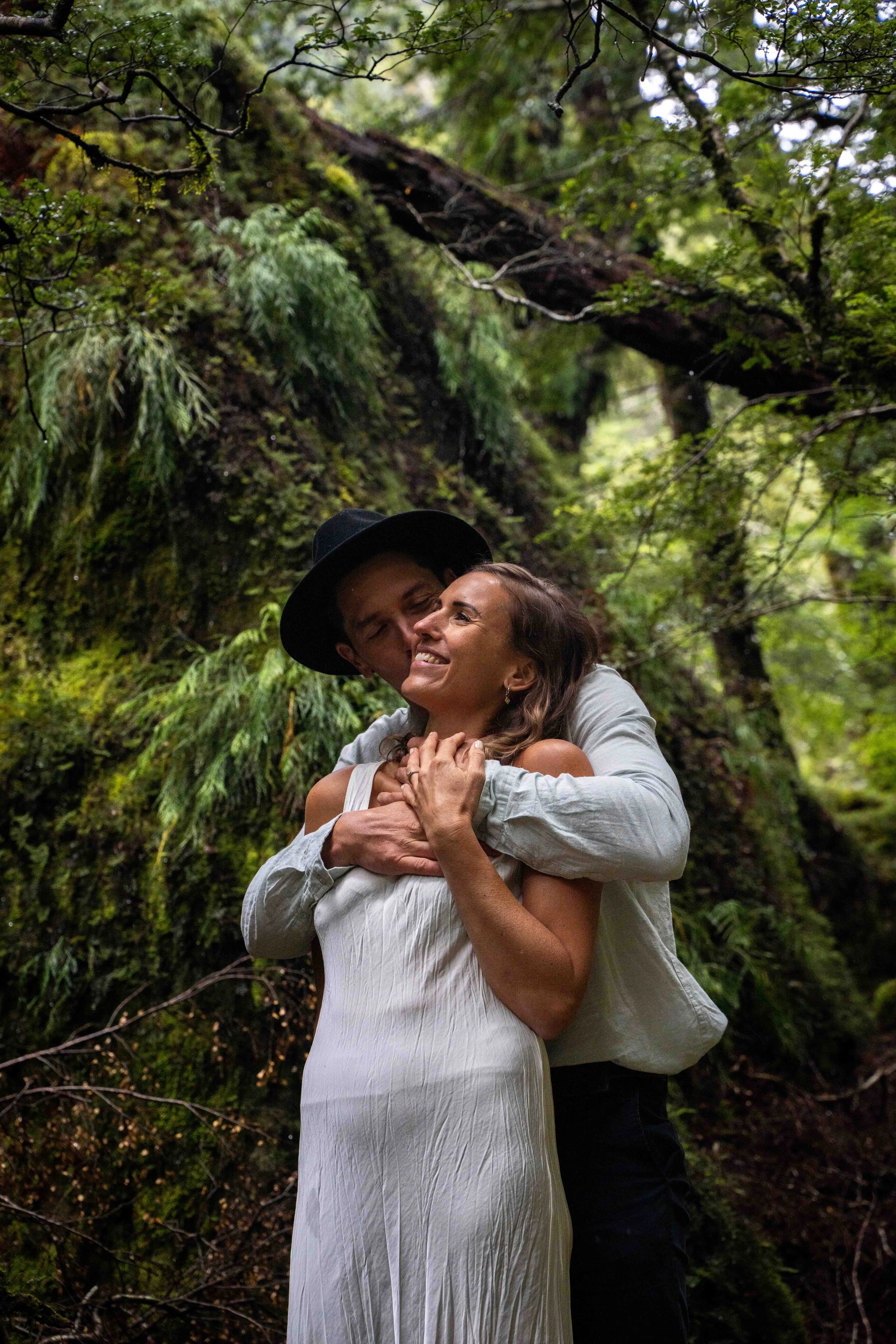 A happy couple embraces in a lush forest setting, surrounded by green foliage and moss-covered trees.