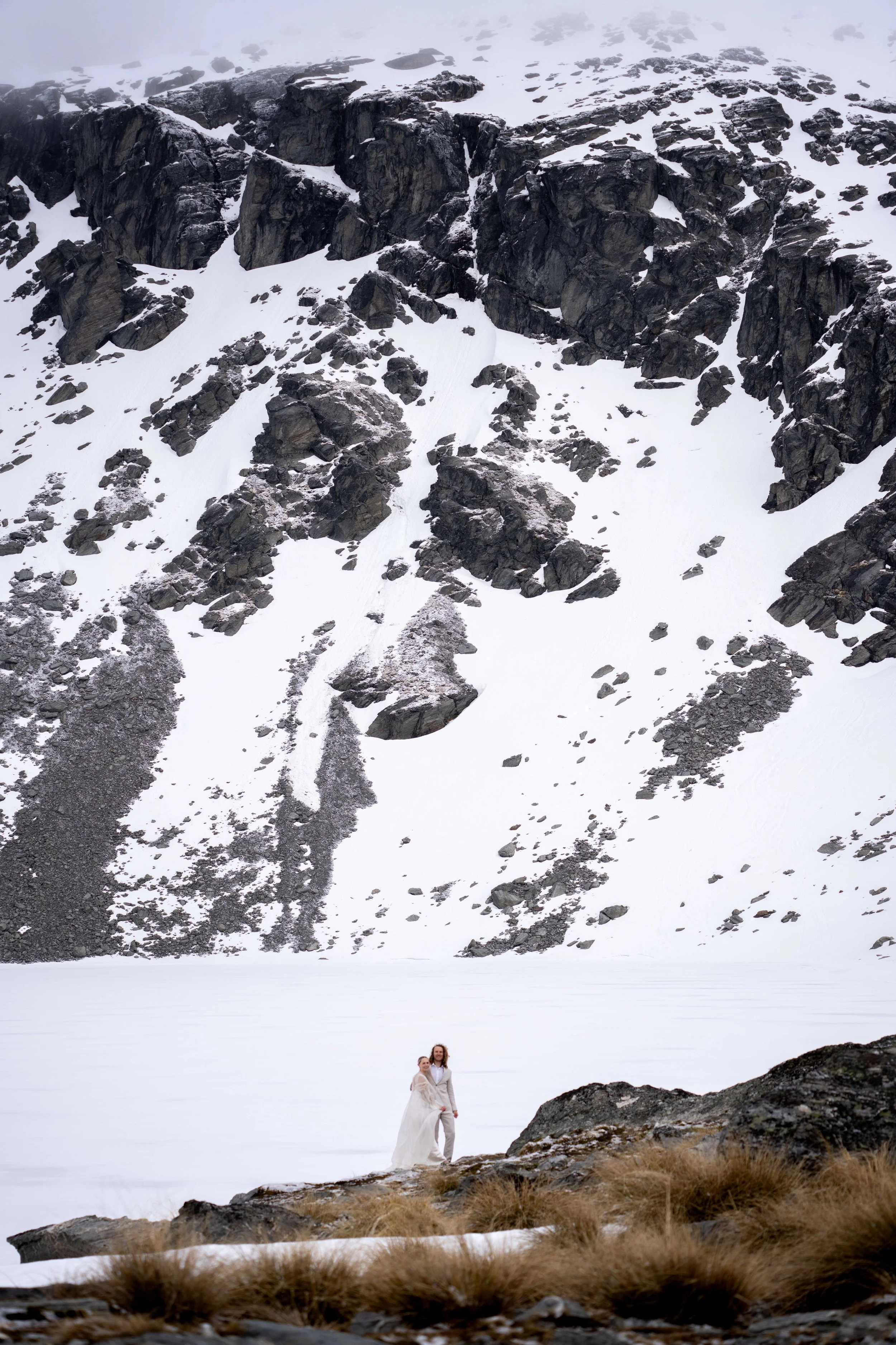 A couple in wedding attire stands on a rocky area in front of a snow-covered lake, with a mountain terrain in the background.