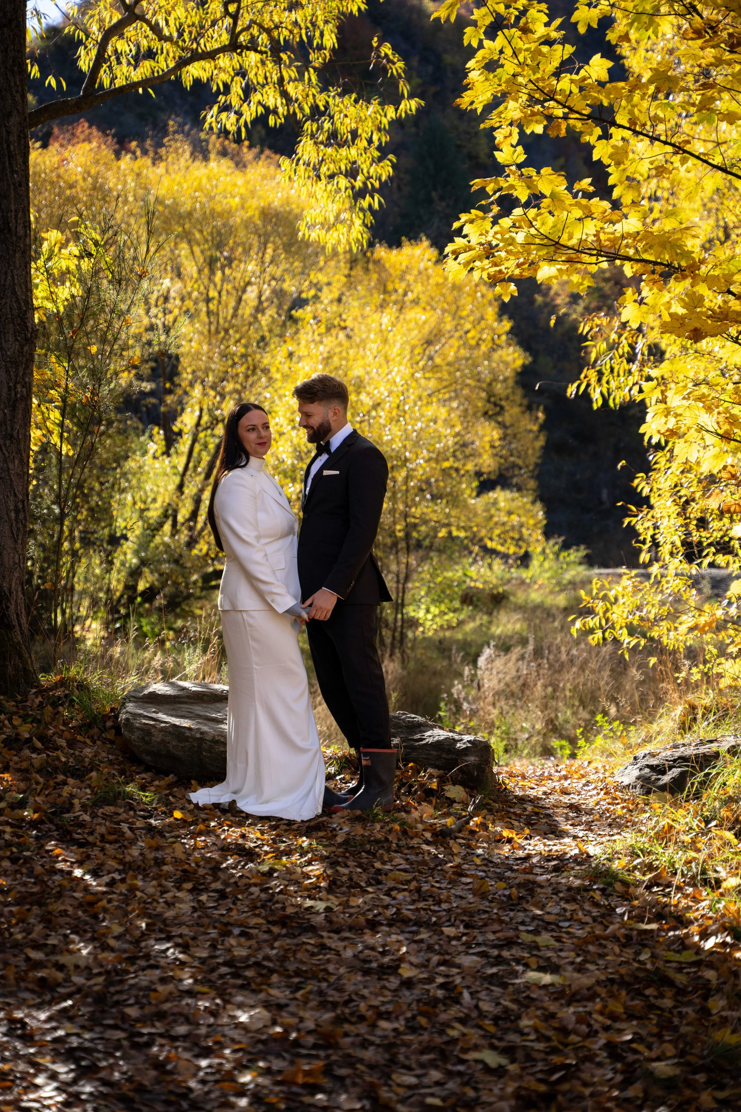 A couple in wedding attire holding hands in an autumn forest with vibrant yellow leaves.