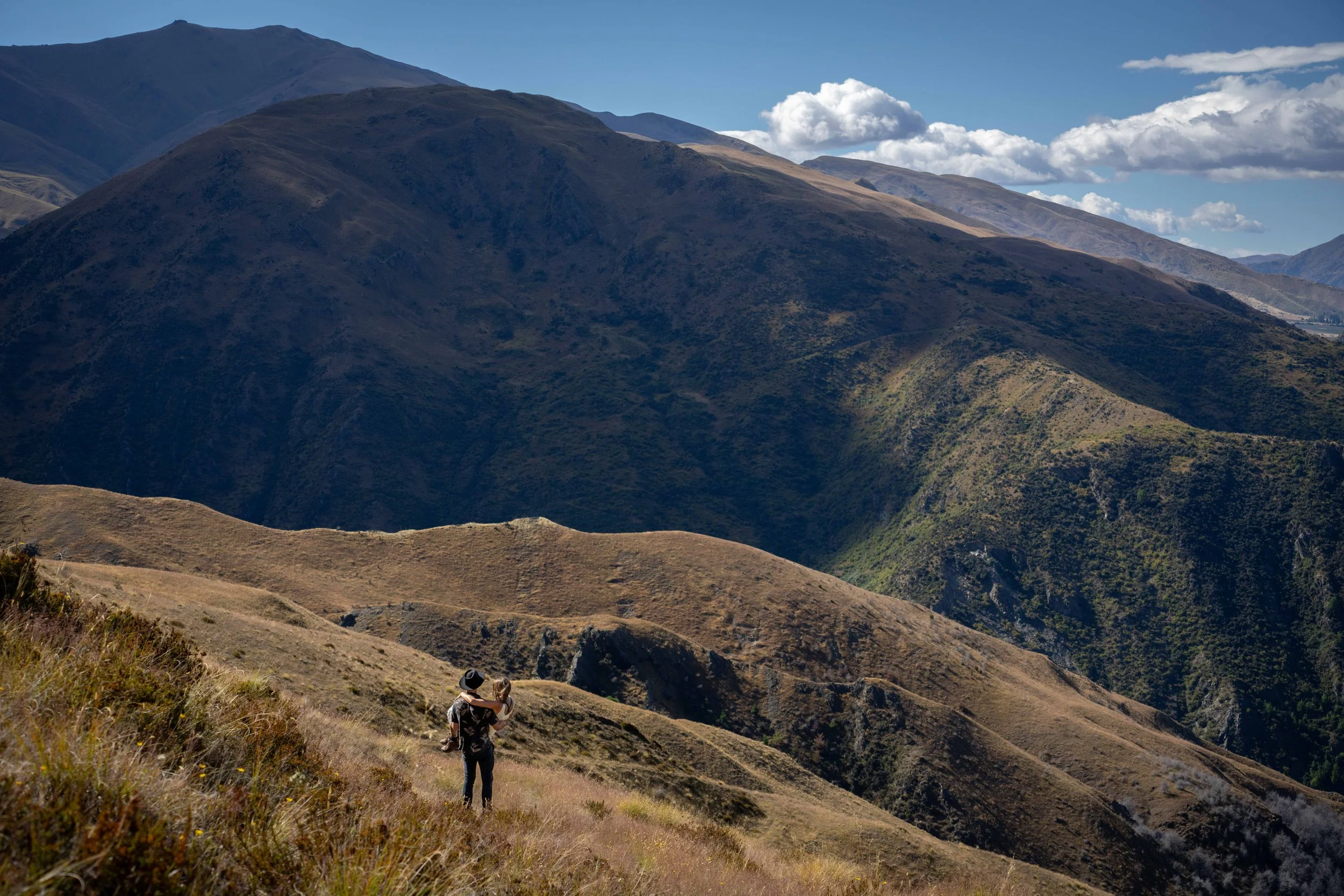 A couple standing on a grassy hillside hiking in a mountainous landscape, with cloudy skies above.