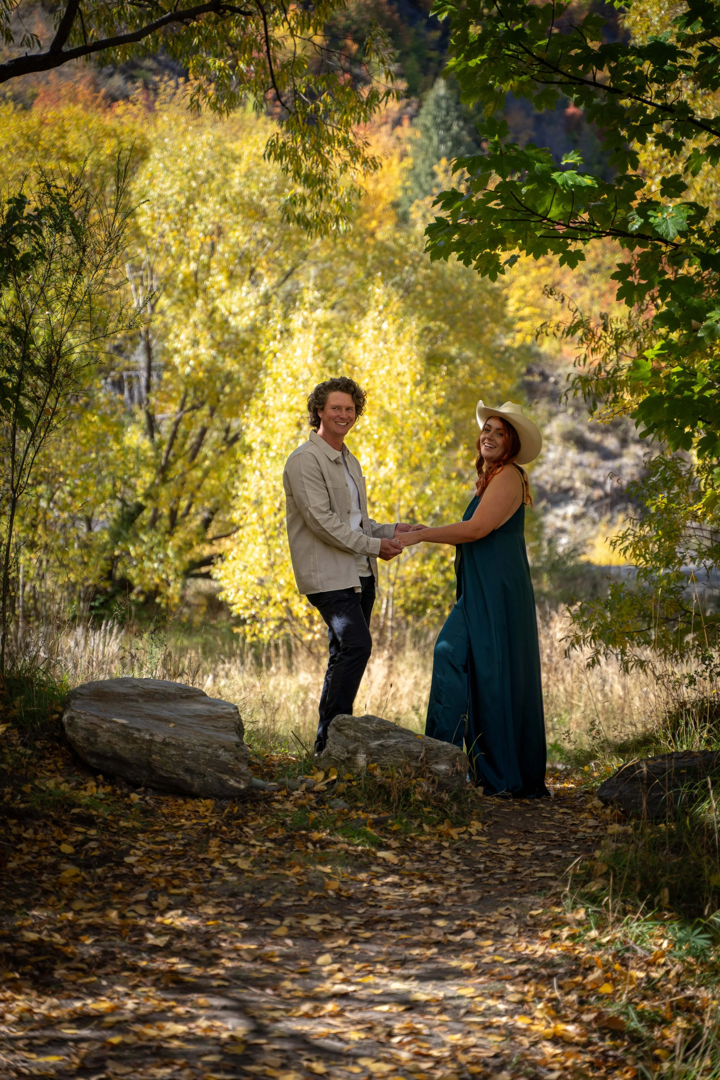 A couple holding hands and smiling in a forest with autumn foliage, rocks, and a dirt path.