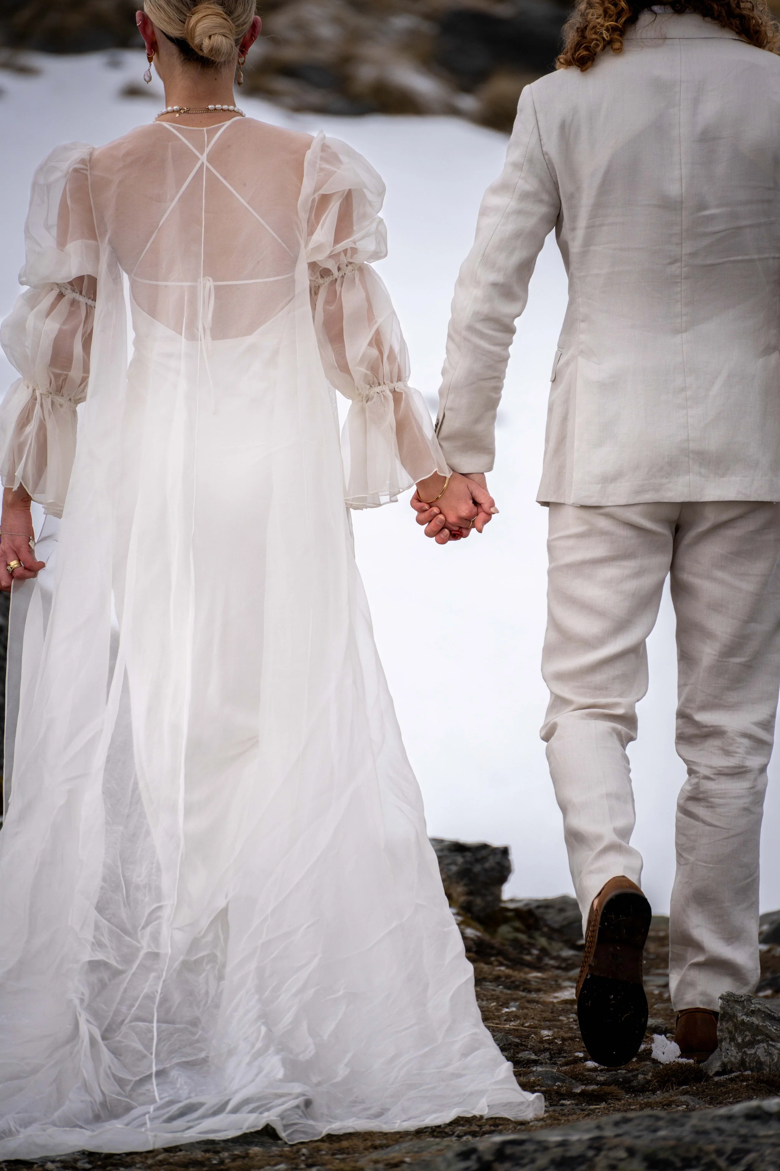 A couple holding hands, dressed in wedding attire, walking outside near the water on a rocky shore.