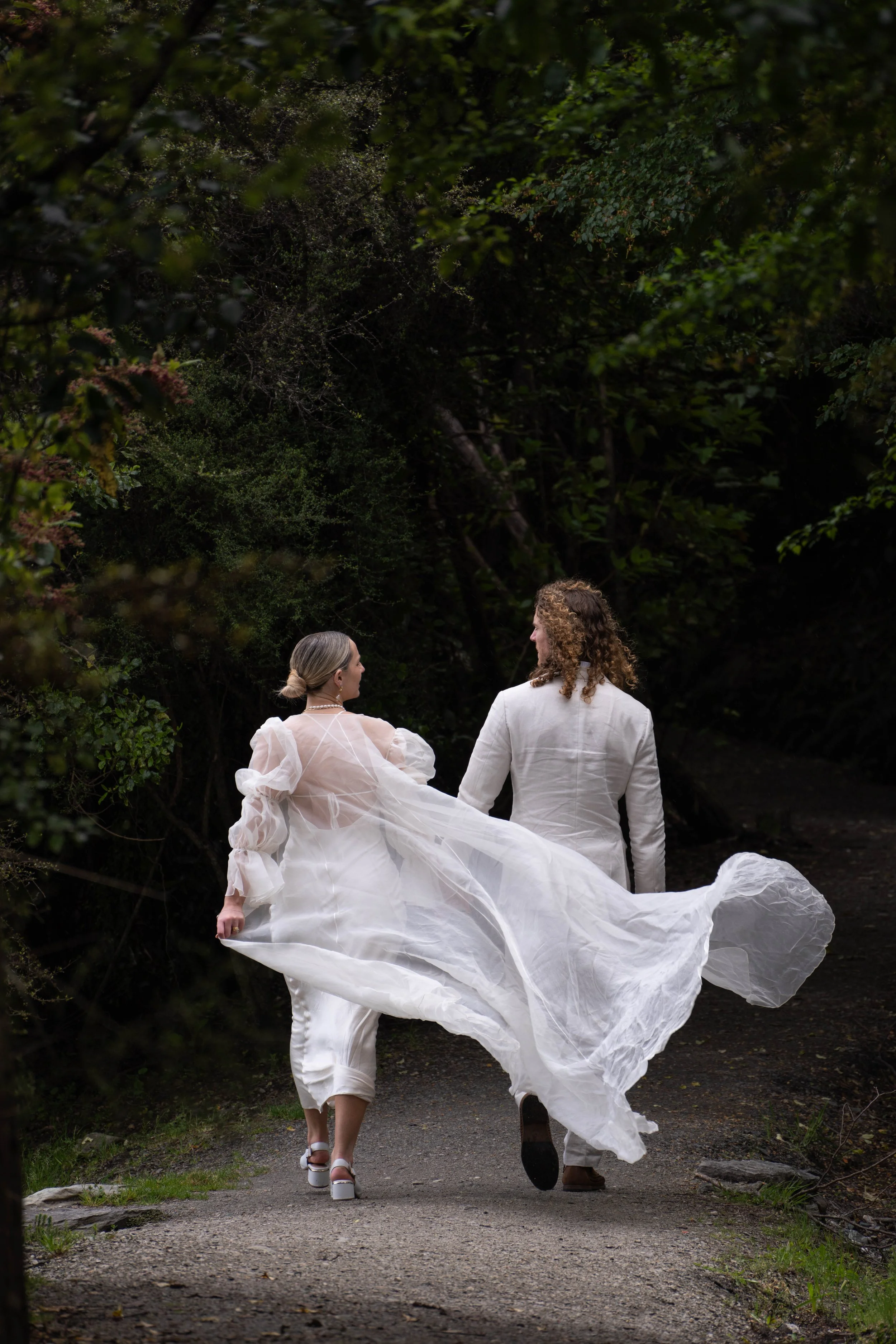 A woman in a white flowing dress and a man in a white suit walk hand in hand on a wooded path.