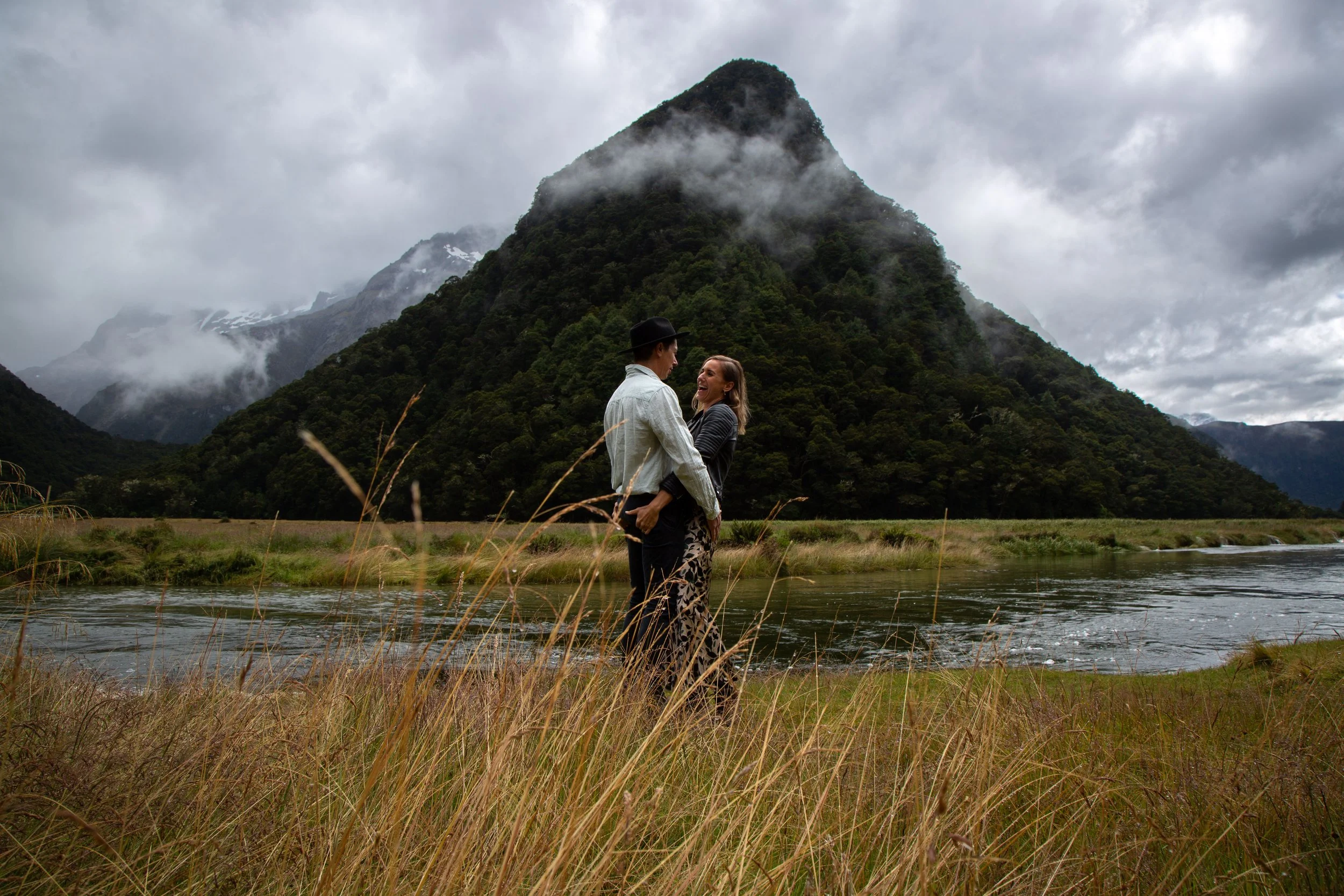 A couple stands holding hands and smiling at each other near a river with a mountainous landscape in the background, during cloudy weather.