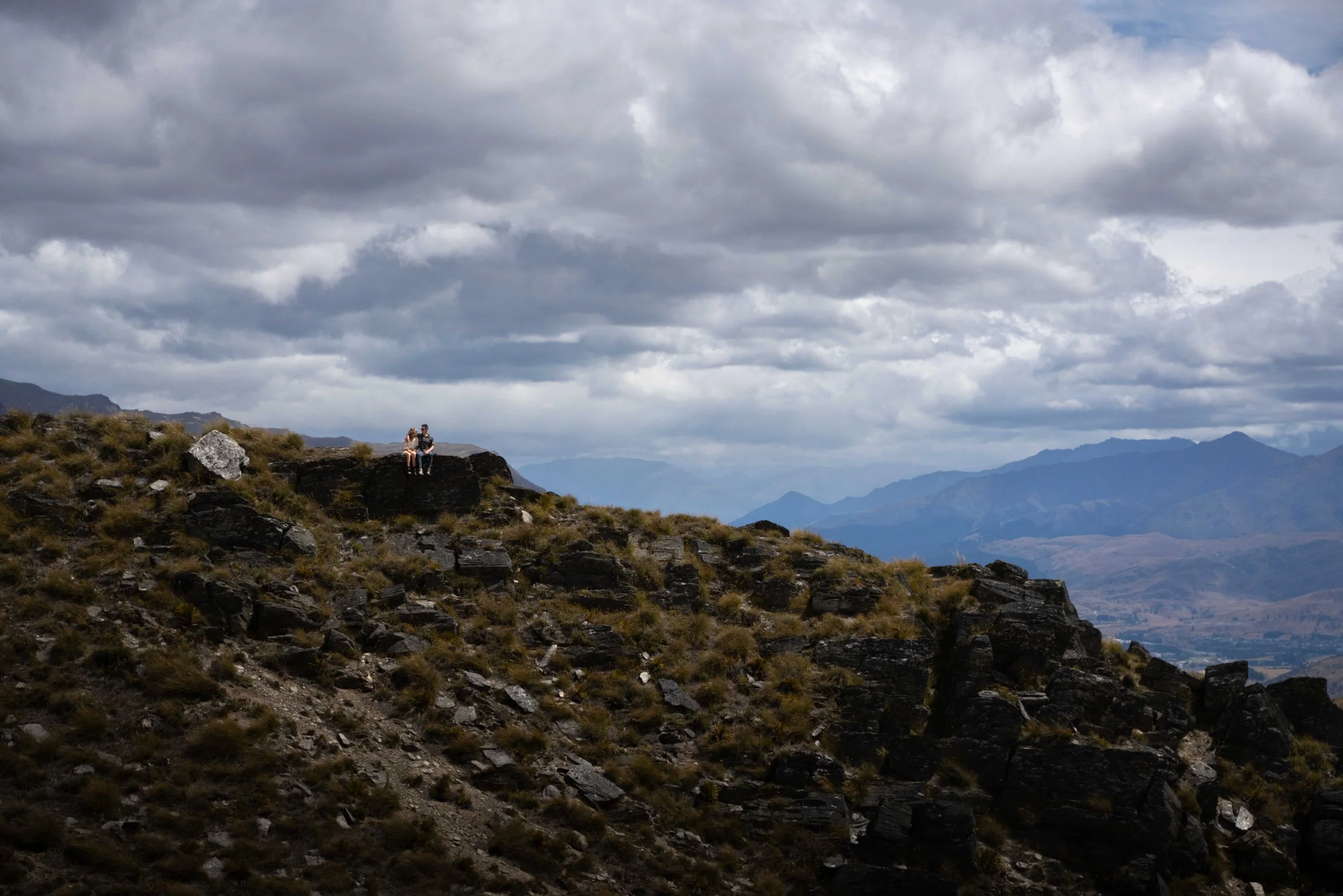 Two people sitting on a large rock on a mountain with cloudy skies and distant mountain ranges in the background.