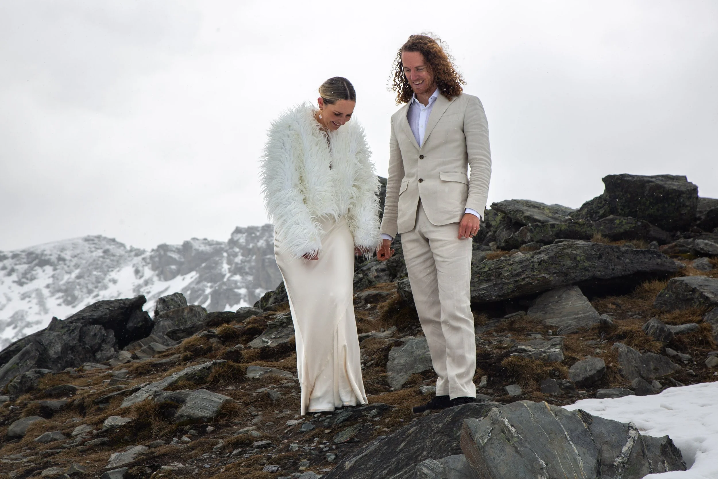 A couple dressed in white wedding attire standing on rocky, snow-covered terrain with mountains in the background.