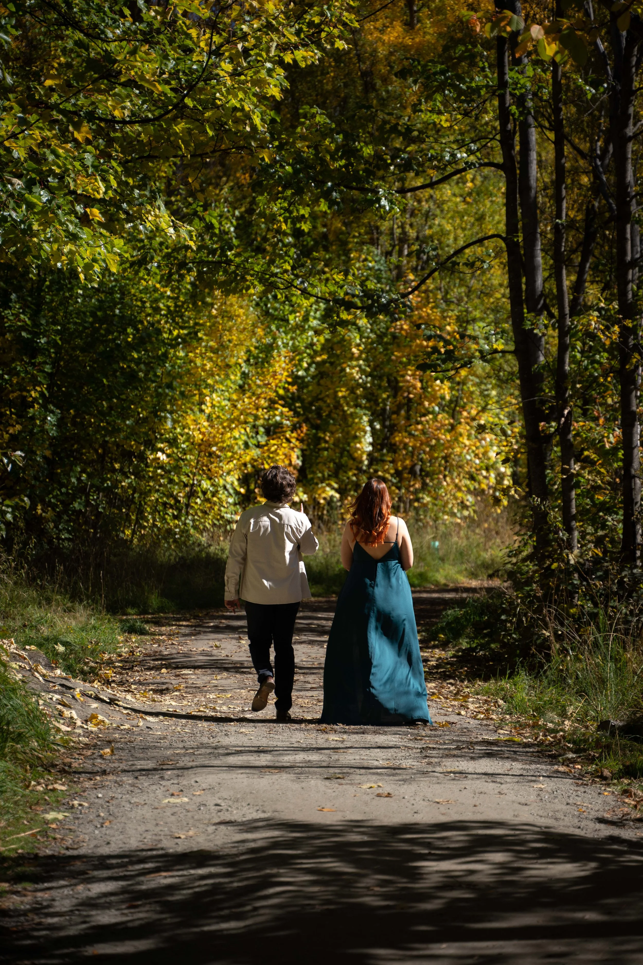 Two people walking along a forest trail surrounded by fall foliage, one in a white jacket and the other in a teal dress.