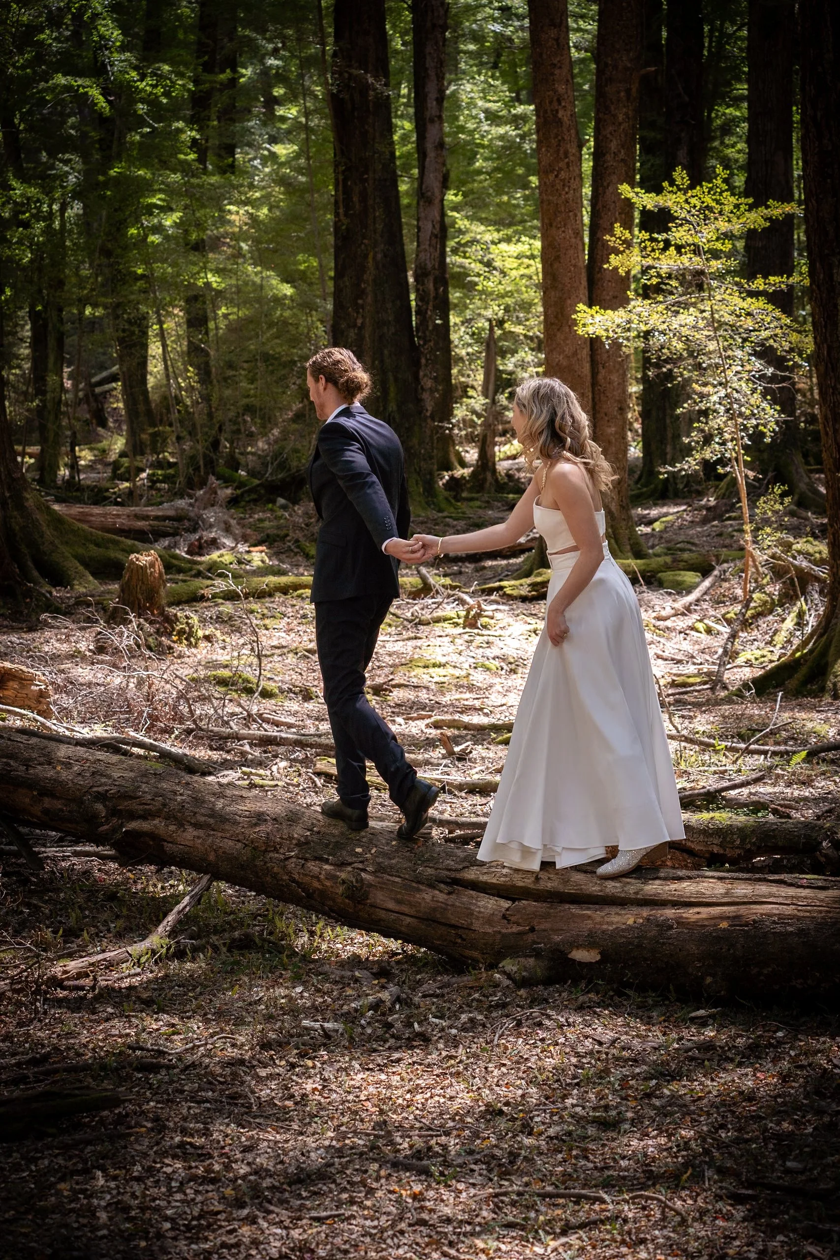 A bride and groom holding hands while walking on a fallen tree in a forest, with sunlight filtering through the trees.