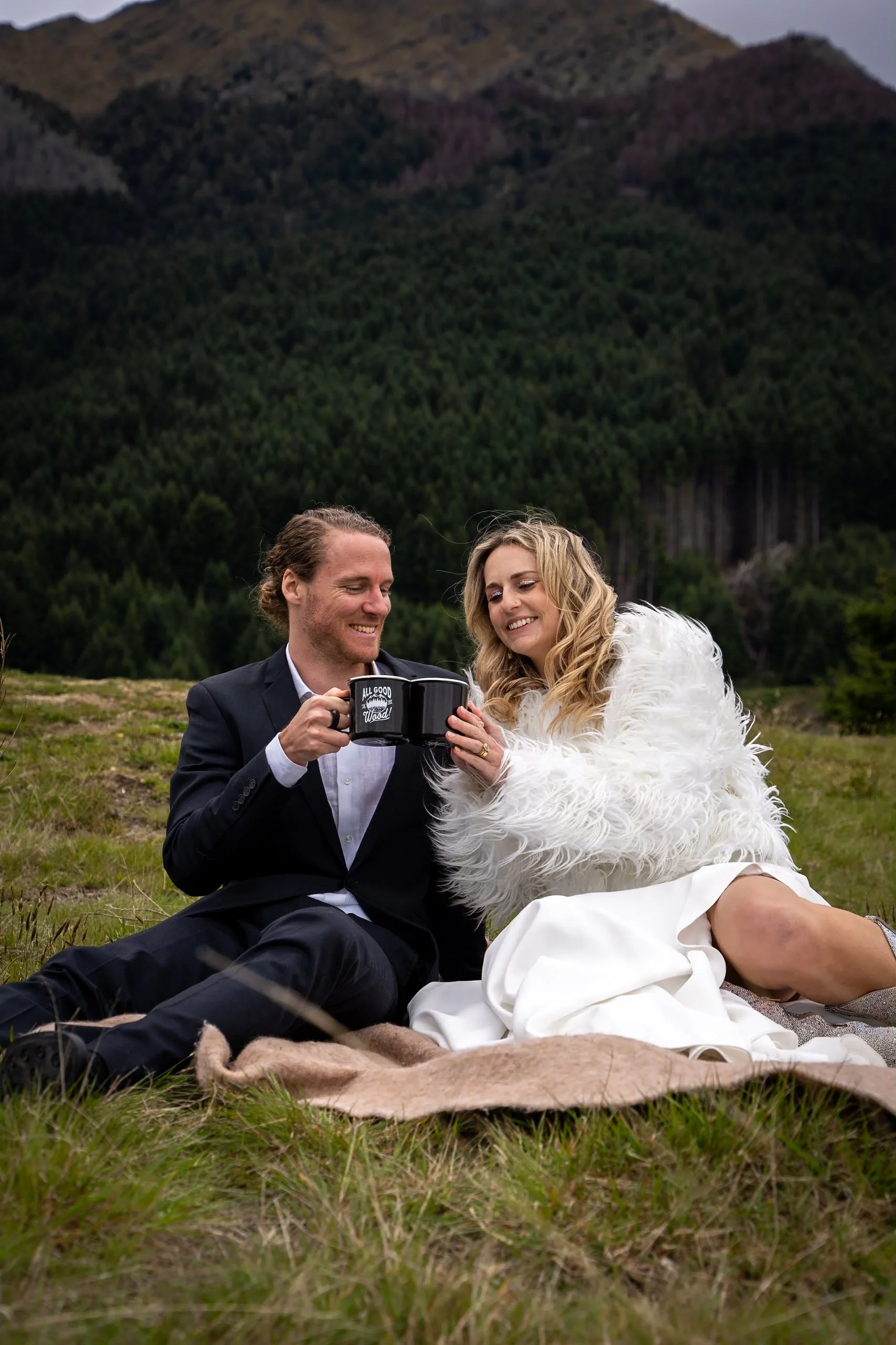 A man and woman sitting on grass outdoors, smiling and looking at a mug they are holding together, with a background of trees and mountains.