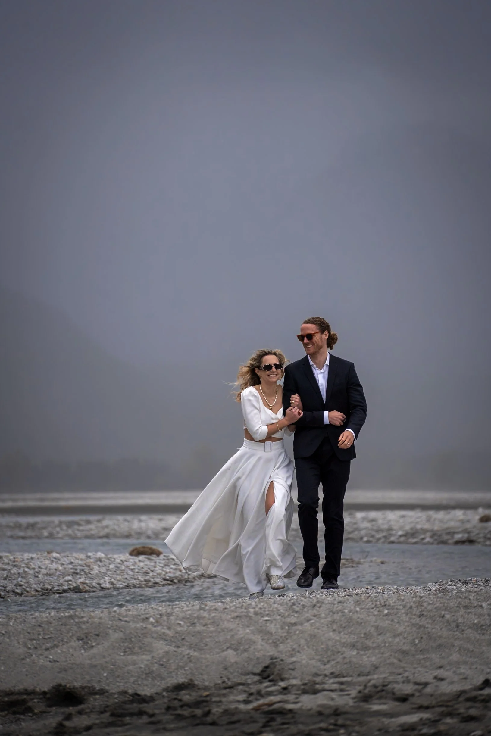 A couple in wedding attire walking on a rocky beach under a cloudy sky, smiling and holding hands.