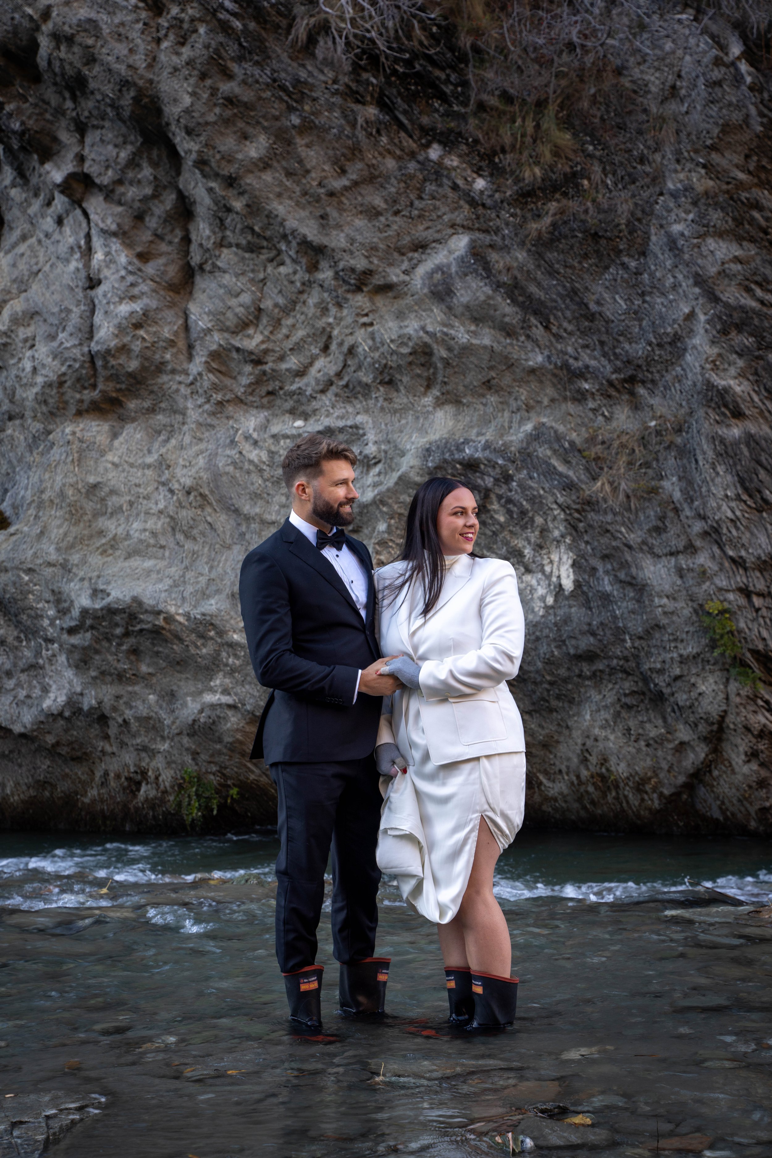 A wedding couple standing in a river at the base of a rocky cliff, dressed in formal attire with waterproof boots, smiling and holding hands.