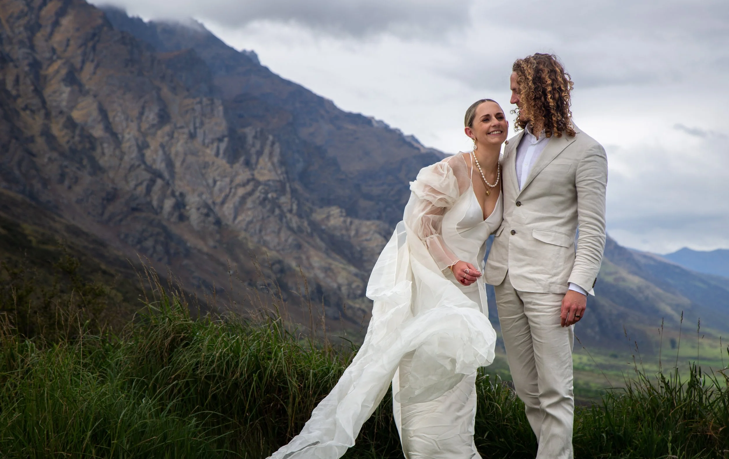 A bride and groom holding hands and smiling outdoors with mountain scenery and cloudy sky in the background.