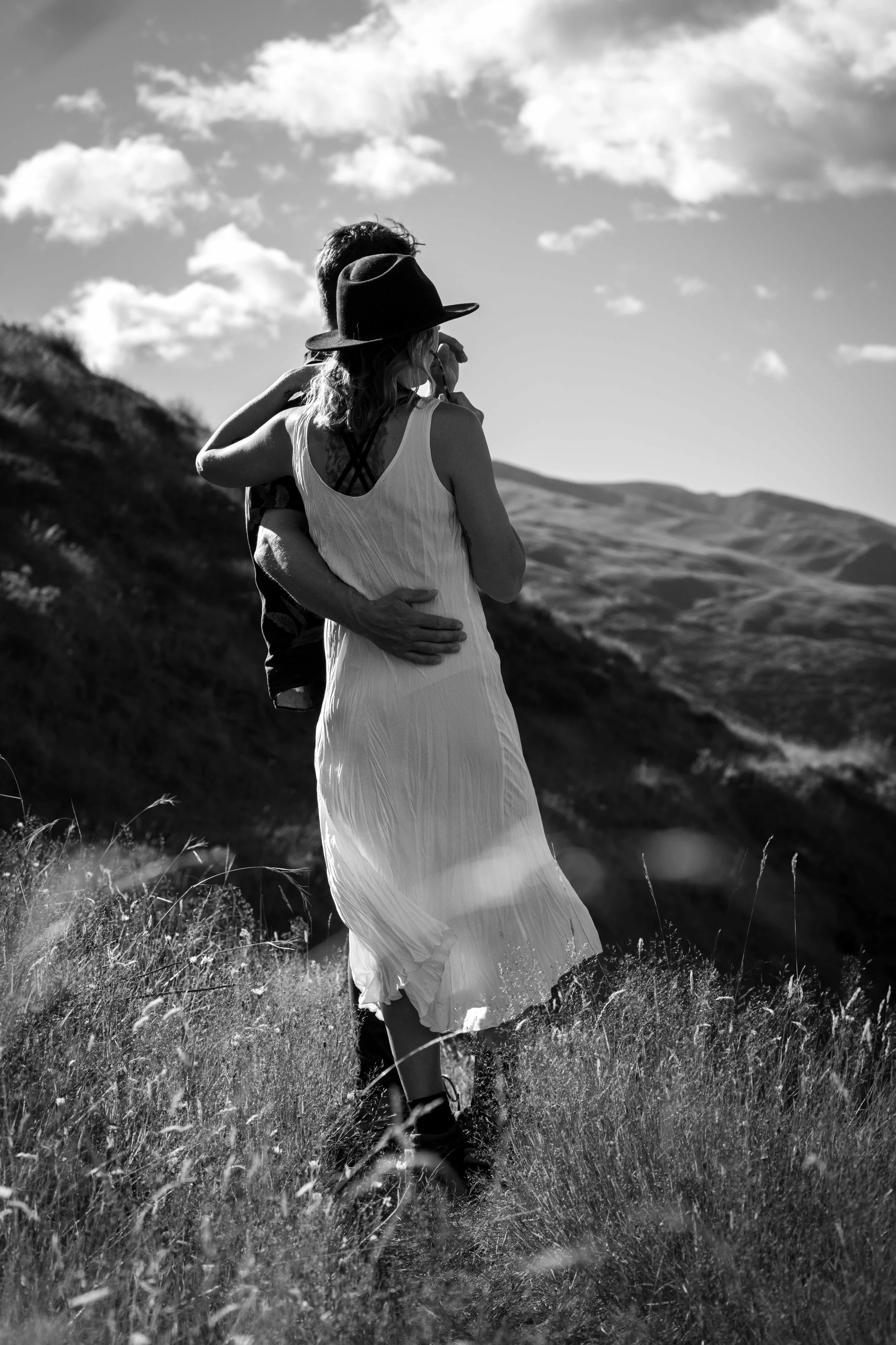 A black-and-white photo of a couple embracing outdoors on a grassy hill, with a landscape of rolling hills and a cloudy sky in the background.