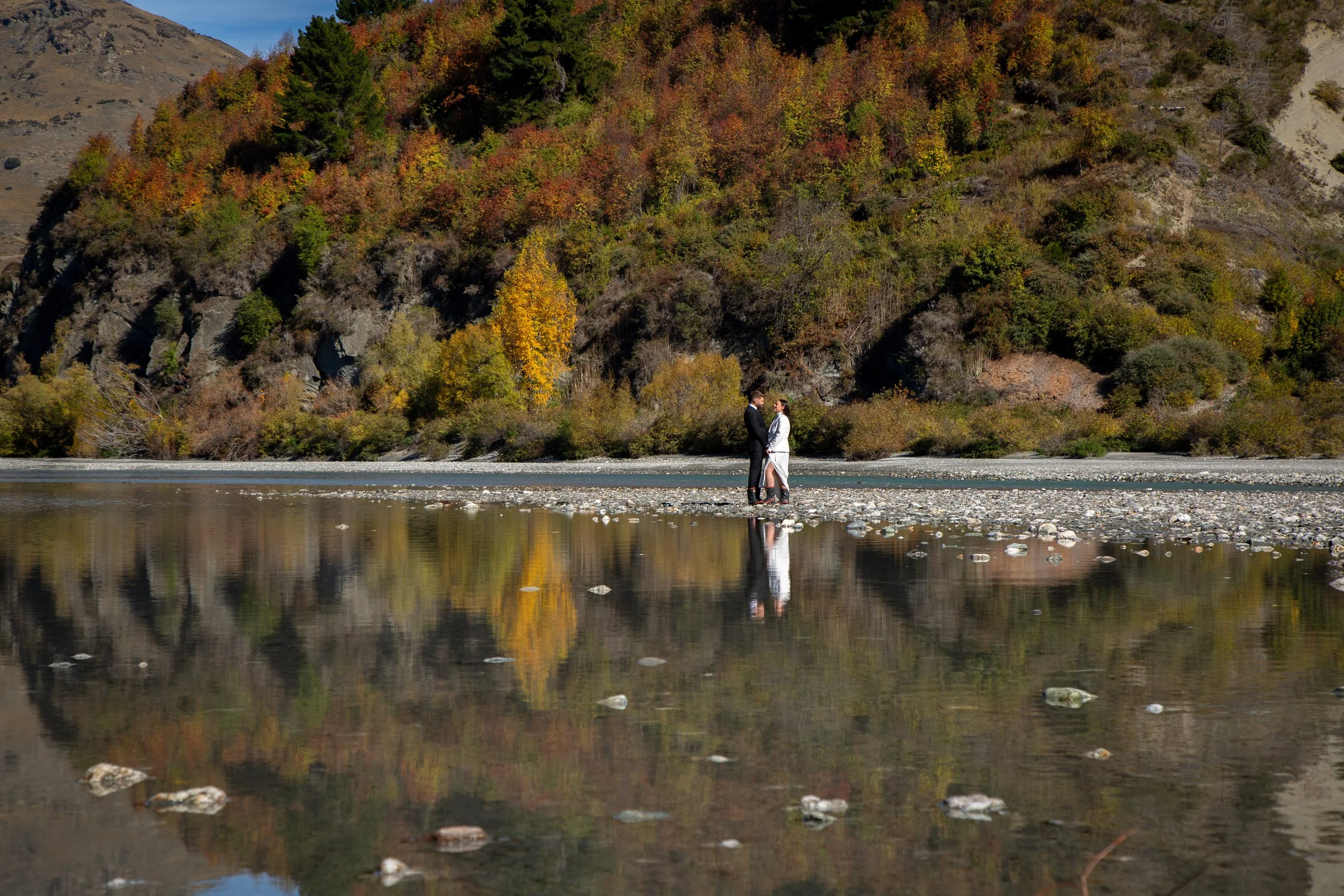 A couple standing on a rocky riverbank, holding hands, with a fall-colored forest and a hillside in the background, and their reflection visible in the water.