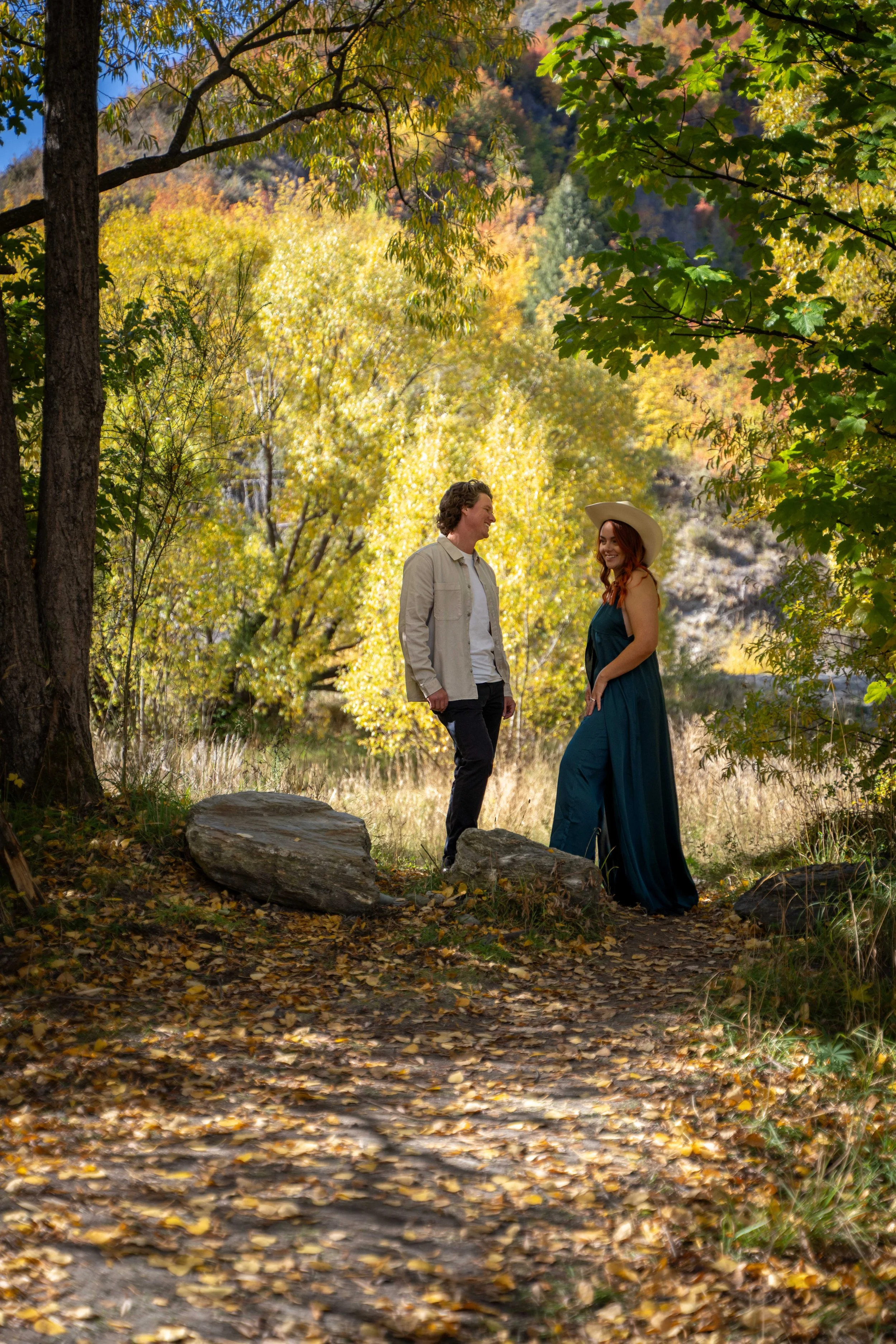 A man and woman standing on a leaf-covered trail in a forest during autumn, smiling at each other, with colorful trees and a hillside in the background.