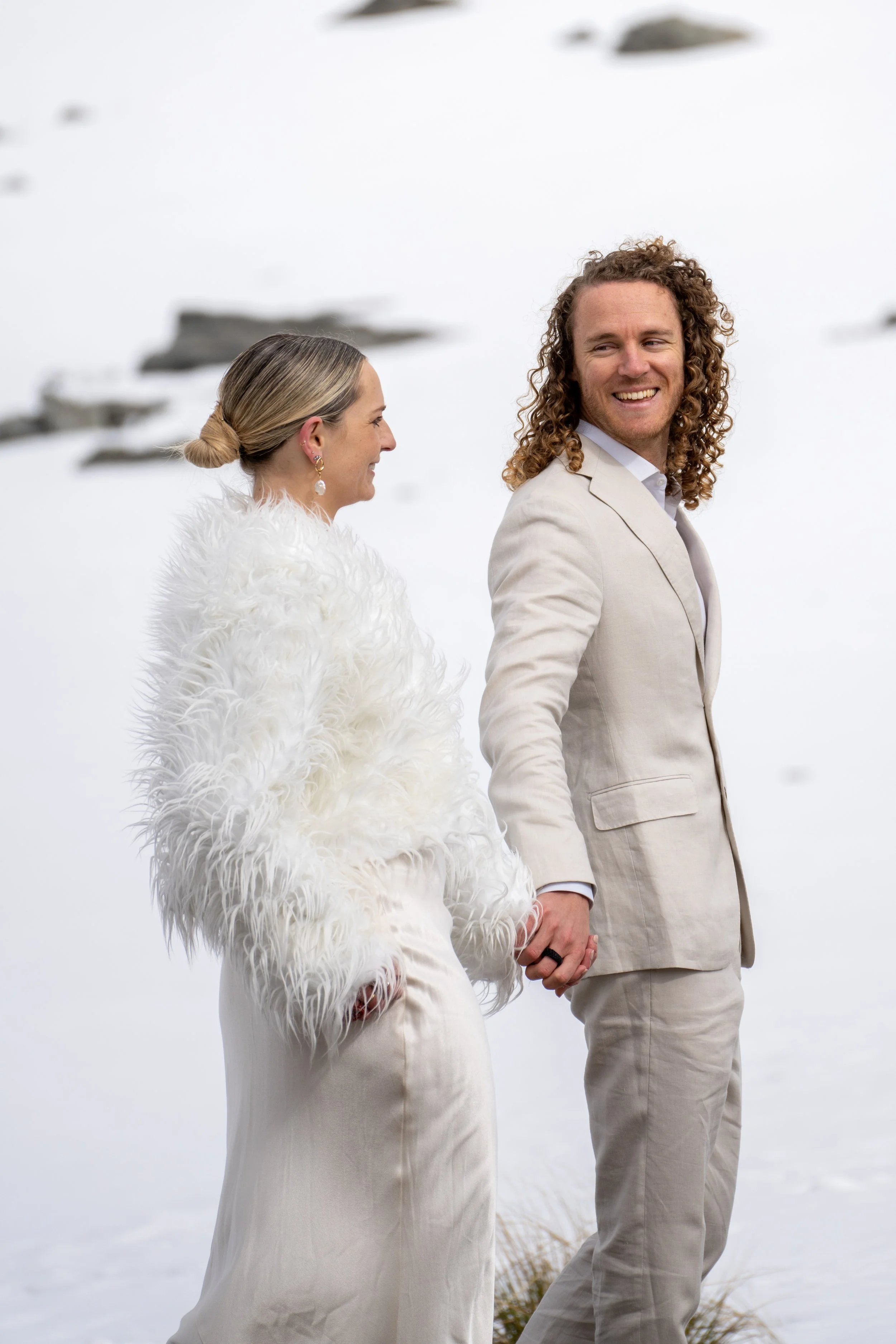 A couple holding hands outdoors in winter, dressed in elegant light-colored clothing, smiling at each other.
