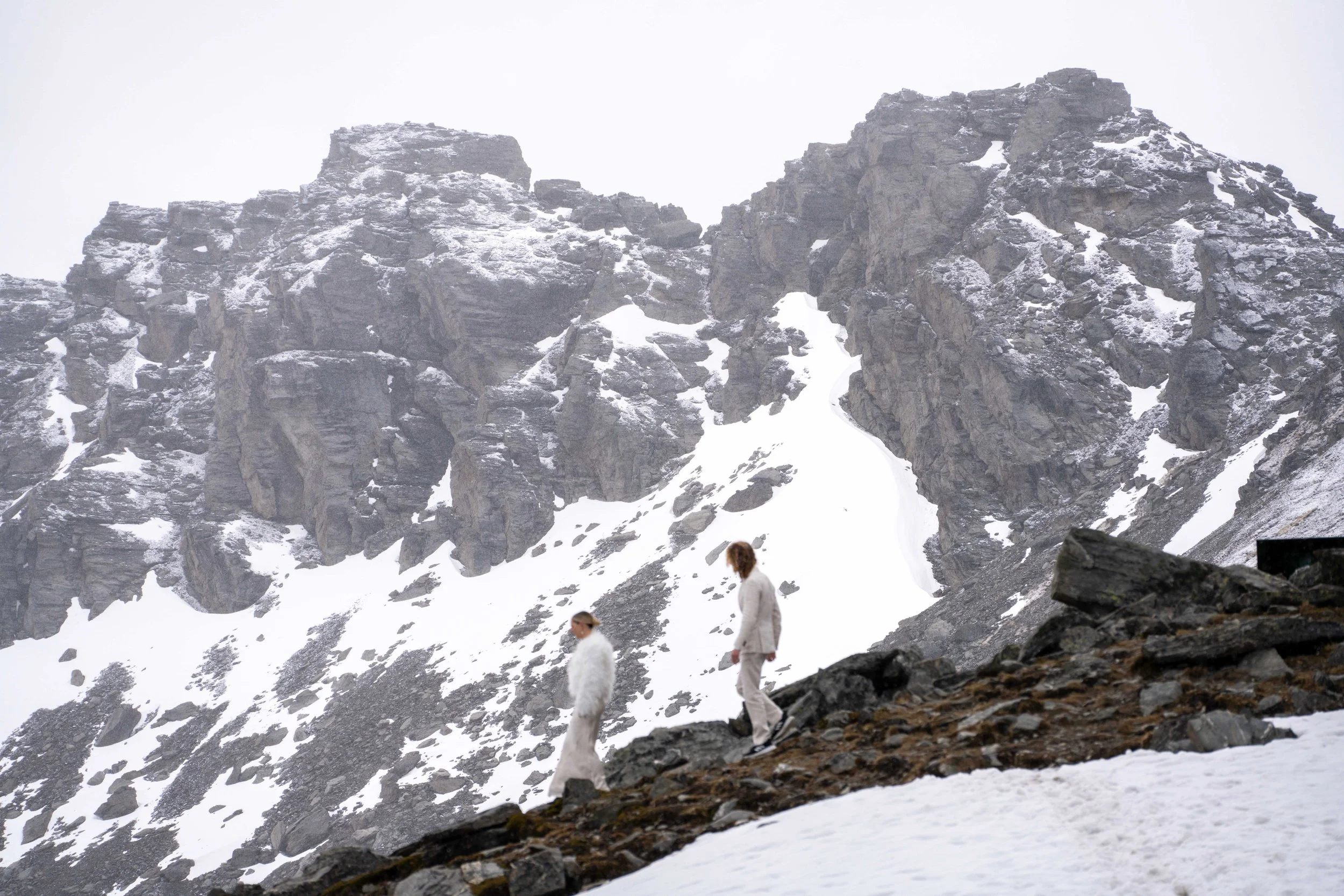 Two people in formal wedding clothing walking down a snow-covered, rocky mountain terrain with steep, rugged, snow-dusted cliffs in the background.