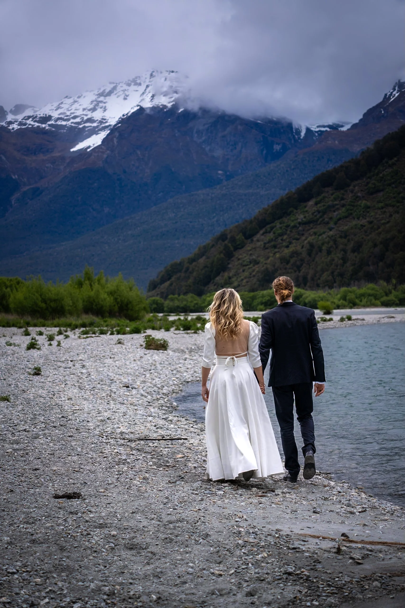 A bride and groom walking hand in hand along a rocky lakeshore with mountains in the background.
