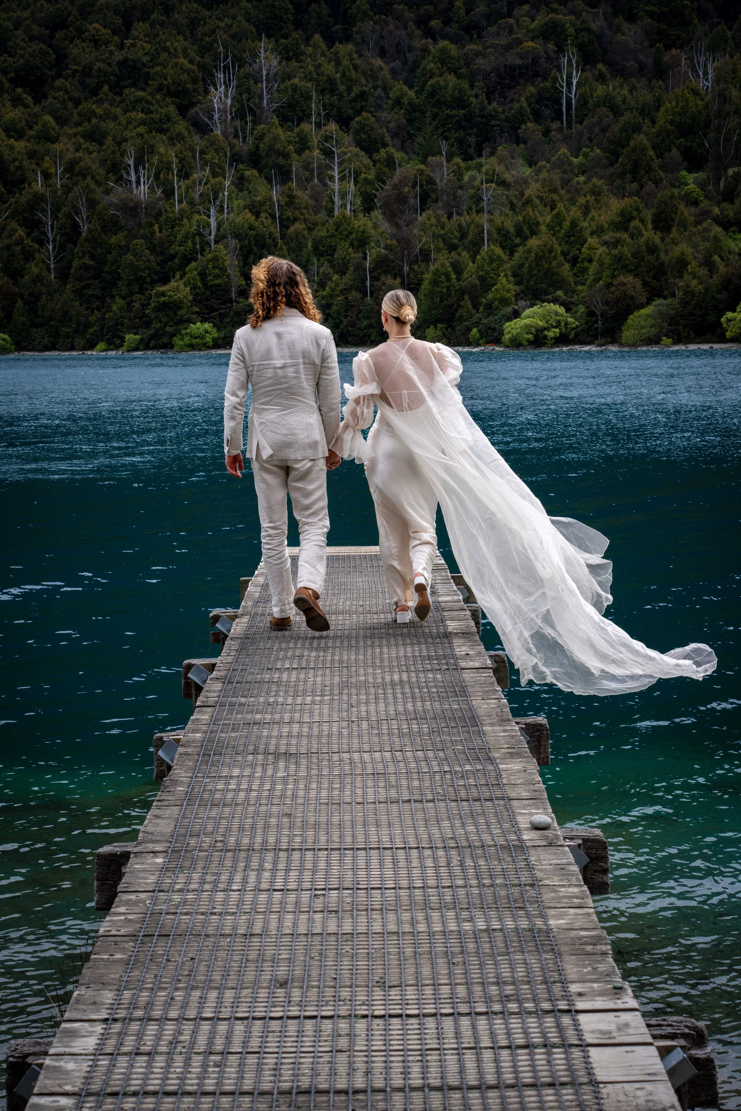 A couple walking hand in hand on a wooden dock by a lake, with a forested hillside in the background. The woman is wearing a flowing white wedding gown, and the man is dressed in a light-colored suit.