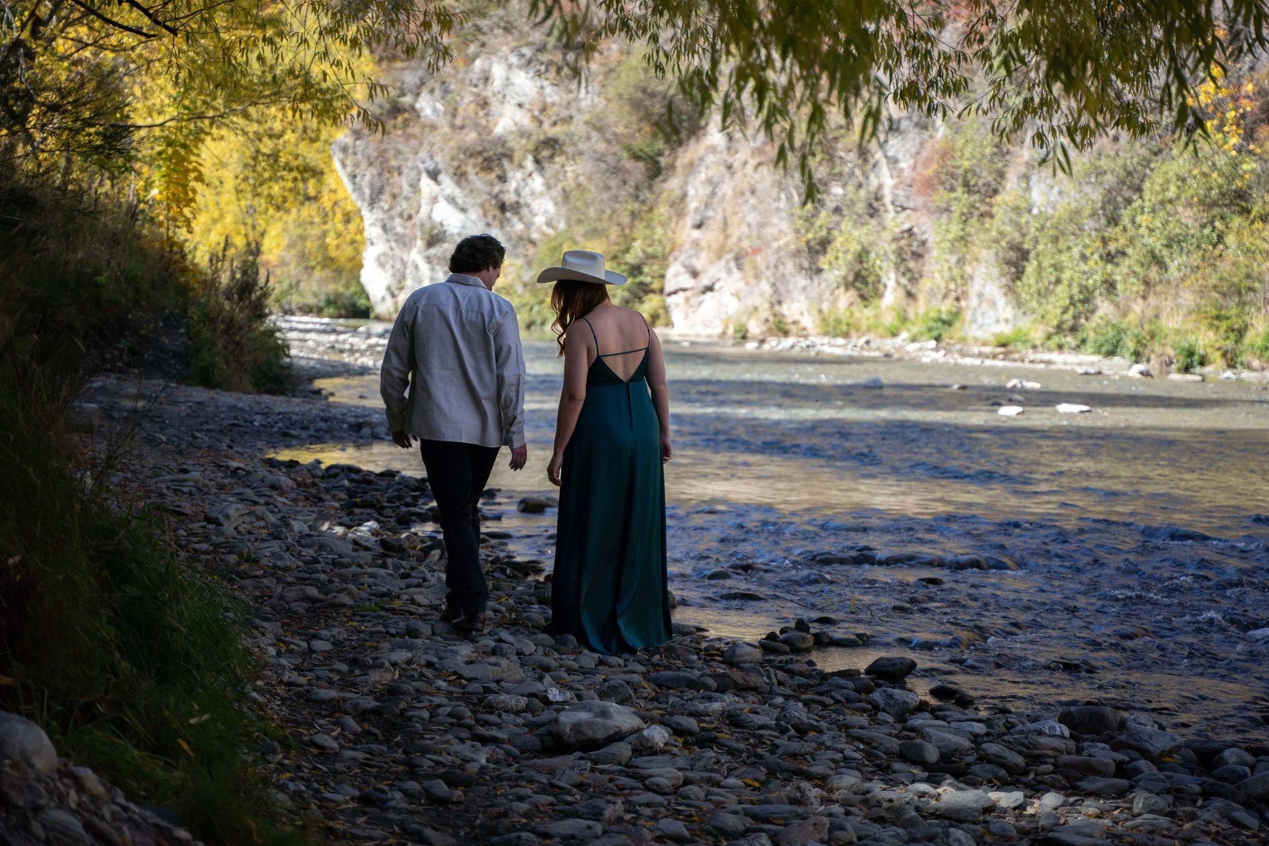 A man and woman walk along a rocky riverbank in a scenic outdoor setting, surrounded by trees and cliffs.