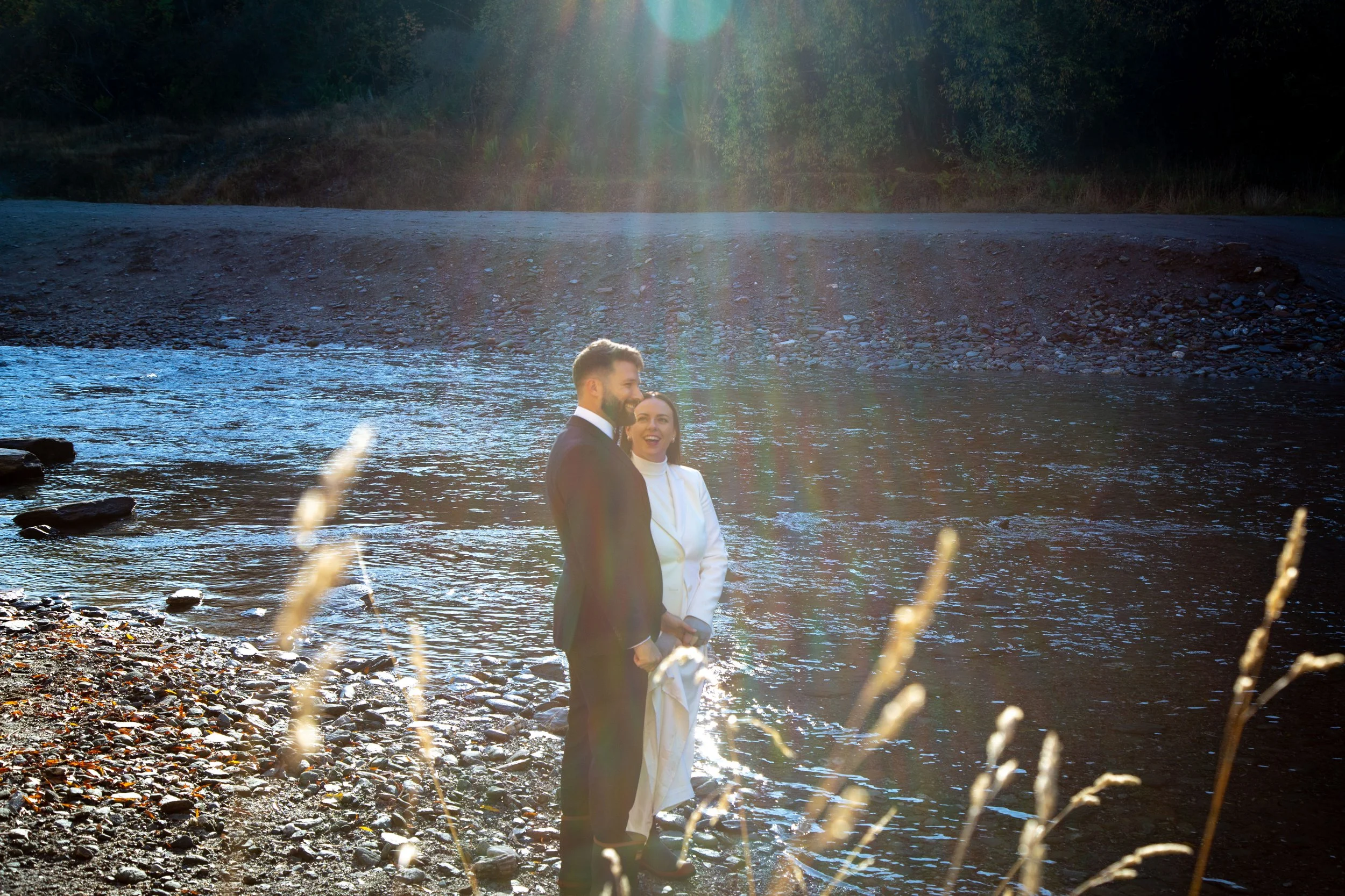 A couple in wedding attire standing by a river at sunset, laughing and holding hands.