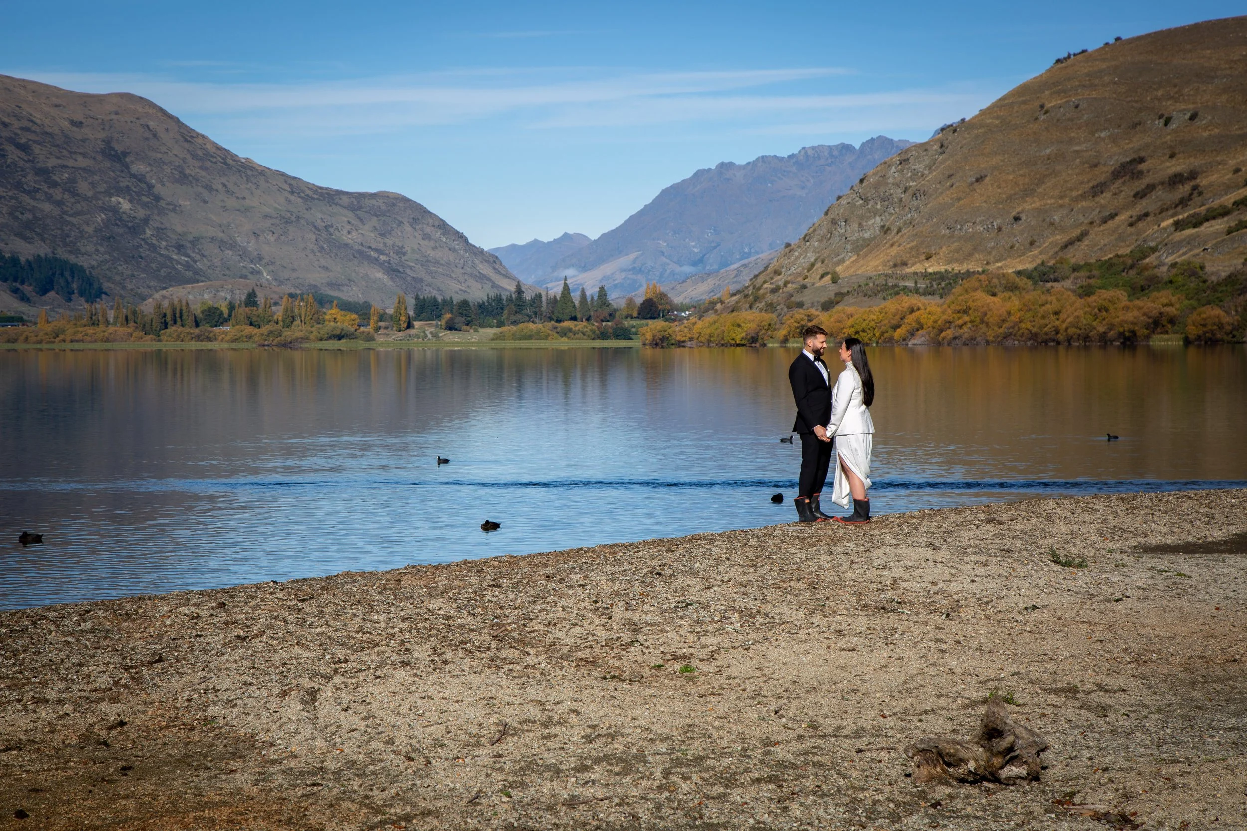 A couple dressed in formal attire standing on a sandy beach by a lake, holding hands and looking at each other, with mountains and trees in the background during daytime.