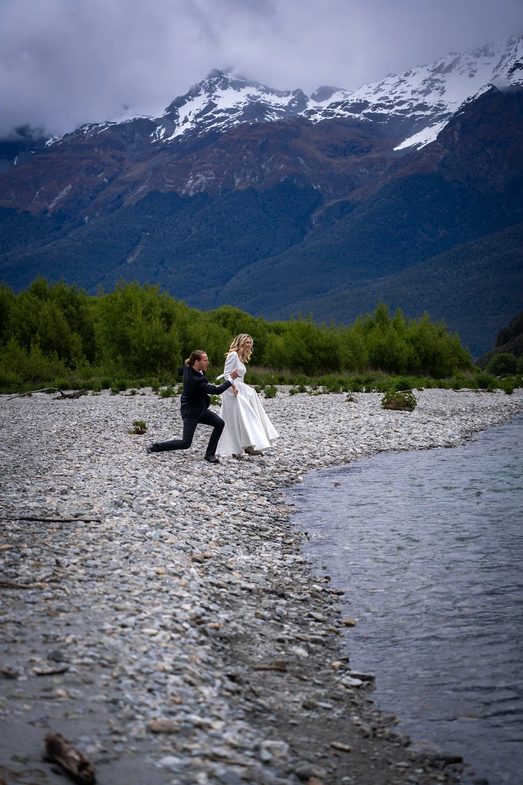 A couple getting married by a river with mountains in the background, the groom kneeling and holding the bride's hand as she walks along the rocky shore.