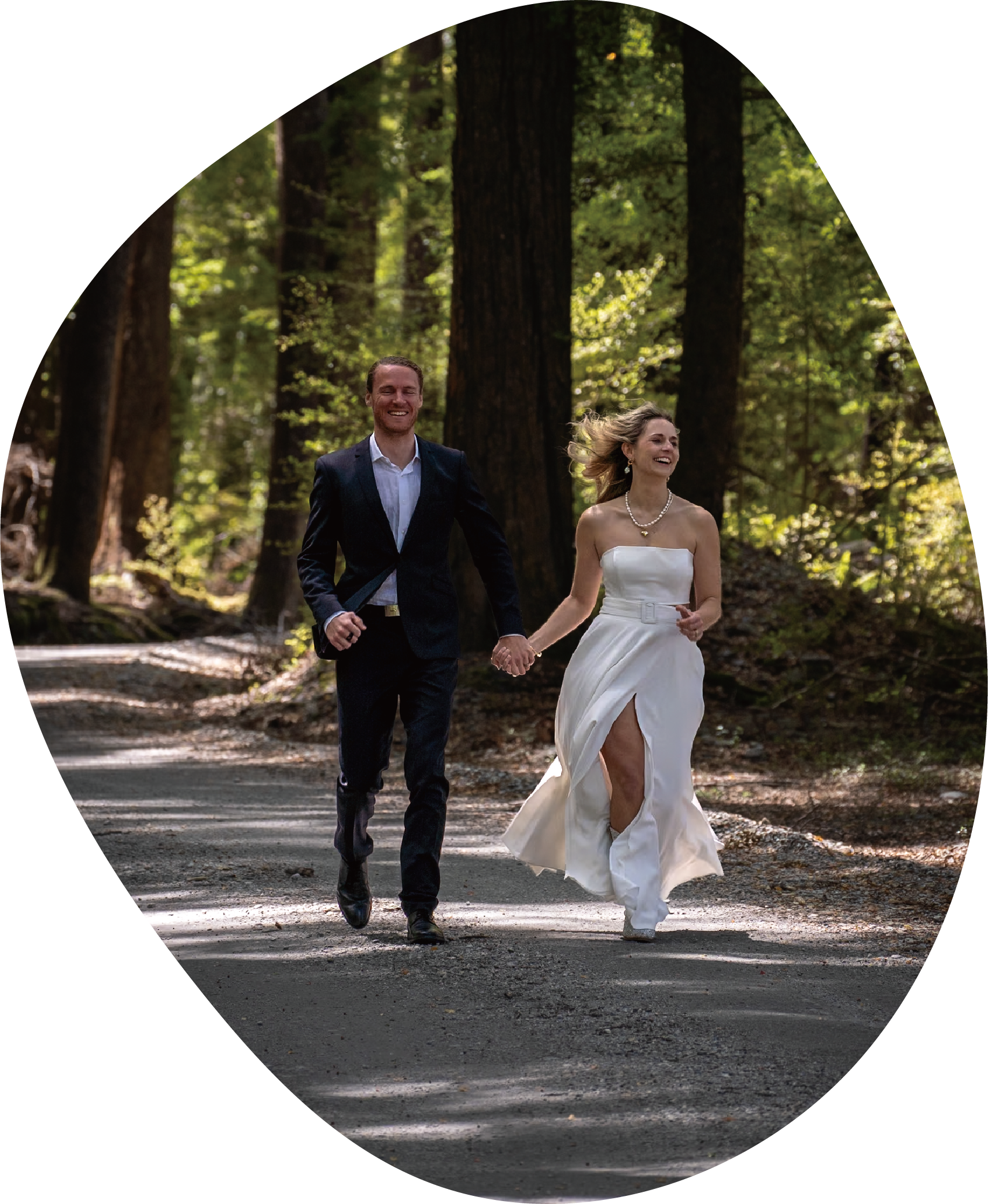 A couple in wedding attire running hand in hand through a wooded forest, with tall trees and sunlight filtering through the leaves.