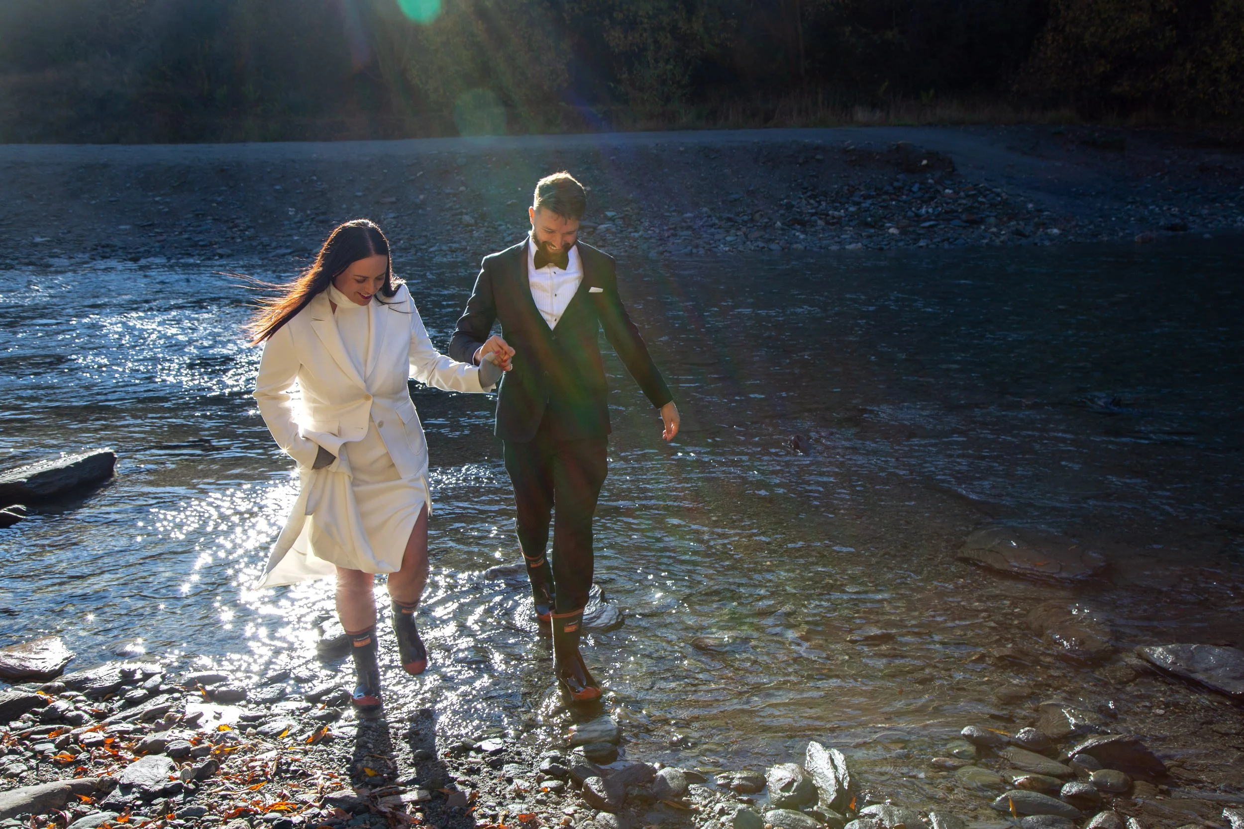 A couple dressed in formal attire laughing and walking hand in hand along a rocky shoreline by a river, with sunlight reflecting on the water surface.