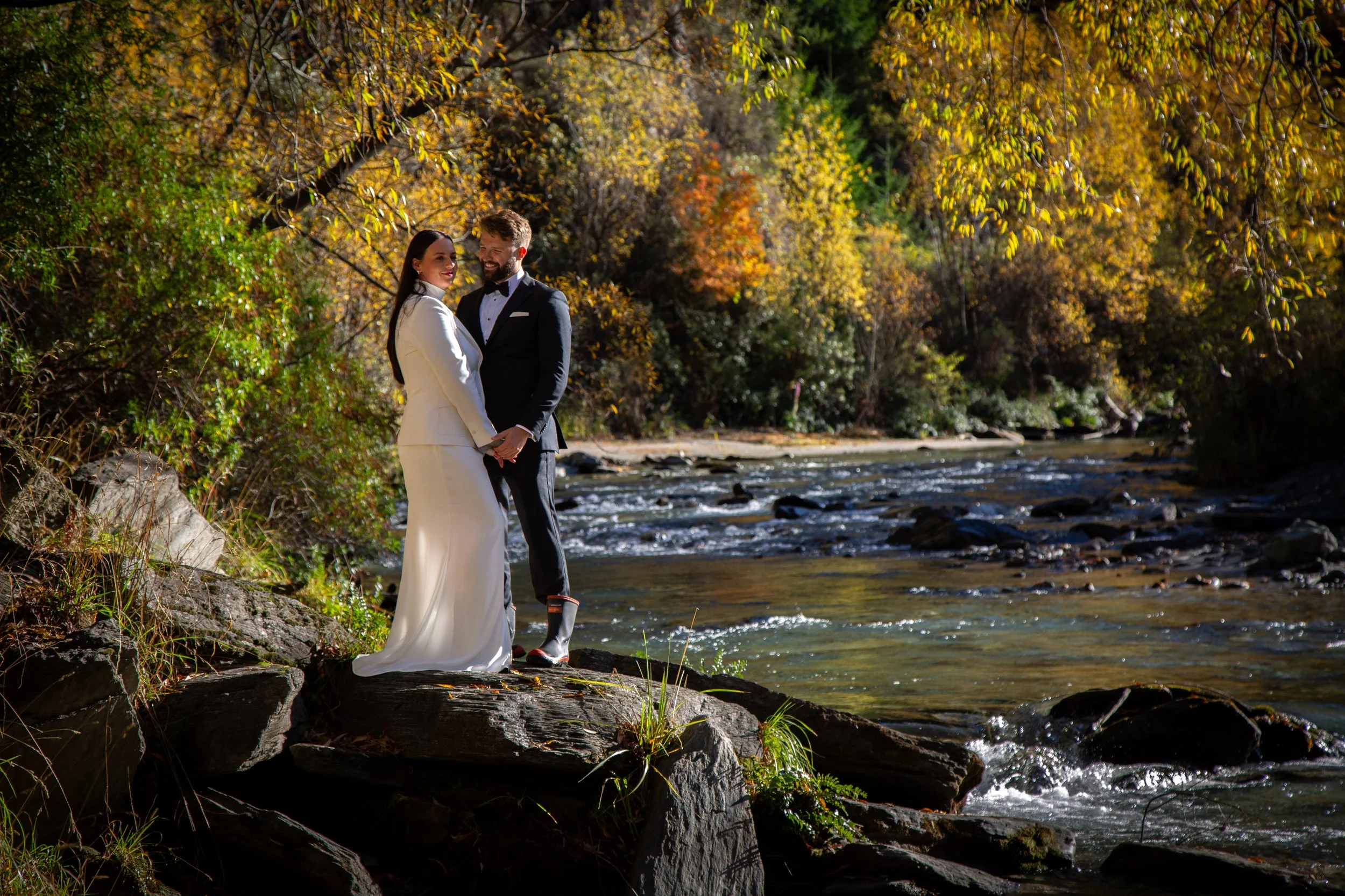 A bride and groom holding hands and smiling at each other on a rocky riverbank surrounded by colorful fall trees.