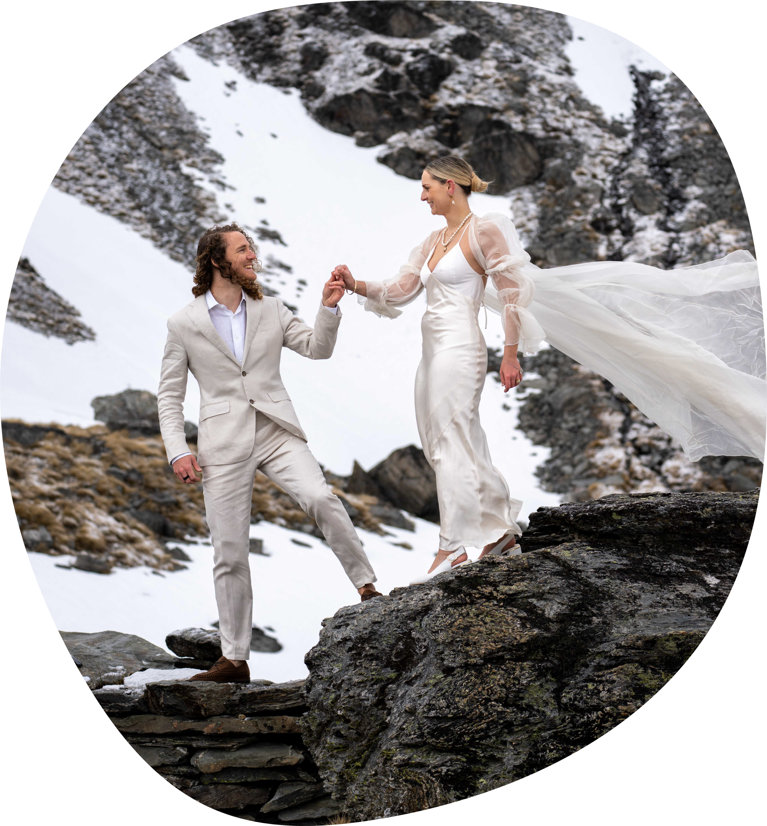 A couple dressed in wedding attire standing on rocks outdoors on a snowy mountain, with the man holding the woman's hand as she steps down.